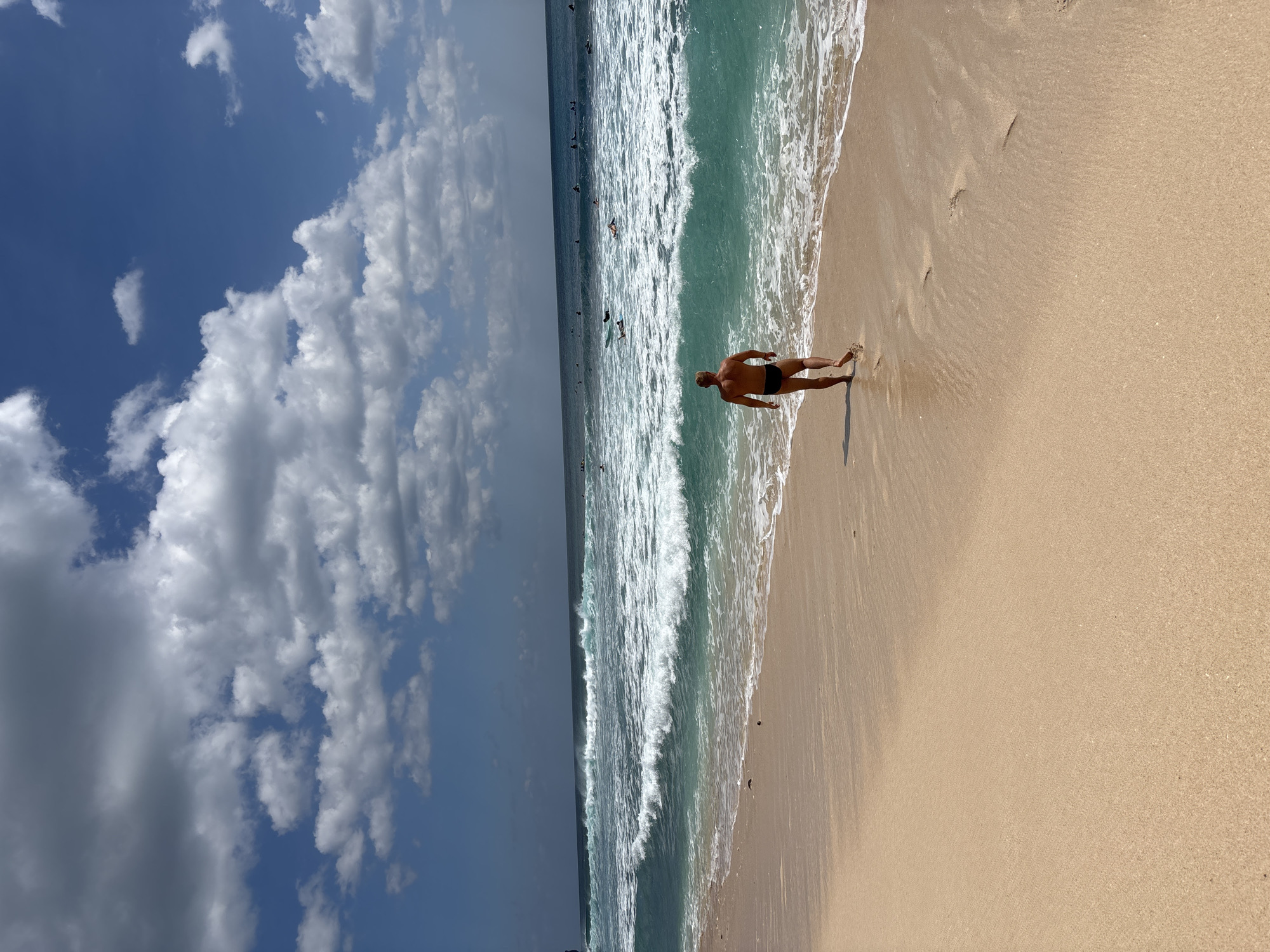 This image captures a daytime beach scene in Kuta Selatan, Indonesia. The upper portion of the frame displays a blue sky with numerous white and grey cumulus clouds. Below the sky, the ocean is visible, transitioning from a light turquoise near the shore to a darker blue further out. Multiple waves are breaking, creating white foam along the coastline. Approximately 15-20 individuals, identified as surfers, are dispersed in the water beyond the immediate breaking waves, some positioned on surfboards. The foreground is composed of light tan sand, with a wet, darker strip along the immediate waterline. A single light-skinned male, with a muscular build, is depicted walking away from the viewer towards the ocean. He wears dark swim trunks, and his footprints are visible in the sand behind him as he approaches the water's edge.