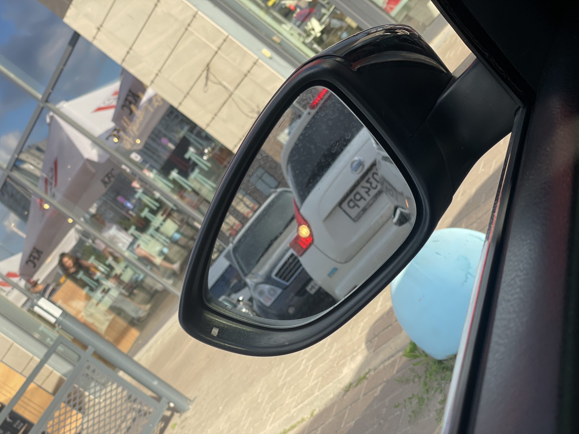 The image is a close-up shot of a car's side-view mirror, taken from inside the vehicle. The mirror reflects the scene behind the car, which appears to be a street with a KFC restaurant. The reflection shows a white car with its brake lights on, a license plate that reads "99 289 BB", and a portion of a dark-colored vehicle in front of it.
In the background, visible through the car window and in the reflection, is a KFC restaurant with large glass windows. Several people are visible inside the restaurant, and there are umbrellas and branding associated with KFC. The building's exterior is modern with a light-colored facade. The ground is paved with brick-like tiles, and there is some greenery visible on the right side of the image, near a large, light-blue inflatable object.
The lighting suggests it is daytime, possibly late afternoon or early evening, given the angle of the light. The weather appears clear. The scene is set in an urban environment, likely a commercial area with parking spaces. The presence of the KFC logo and the license plate format provide location cues. No specific emotions are evident, but the overall impression is that of a typical day in a city.