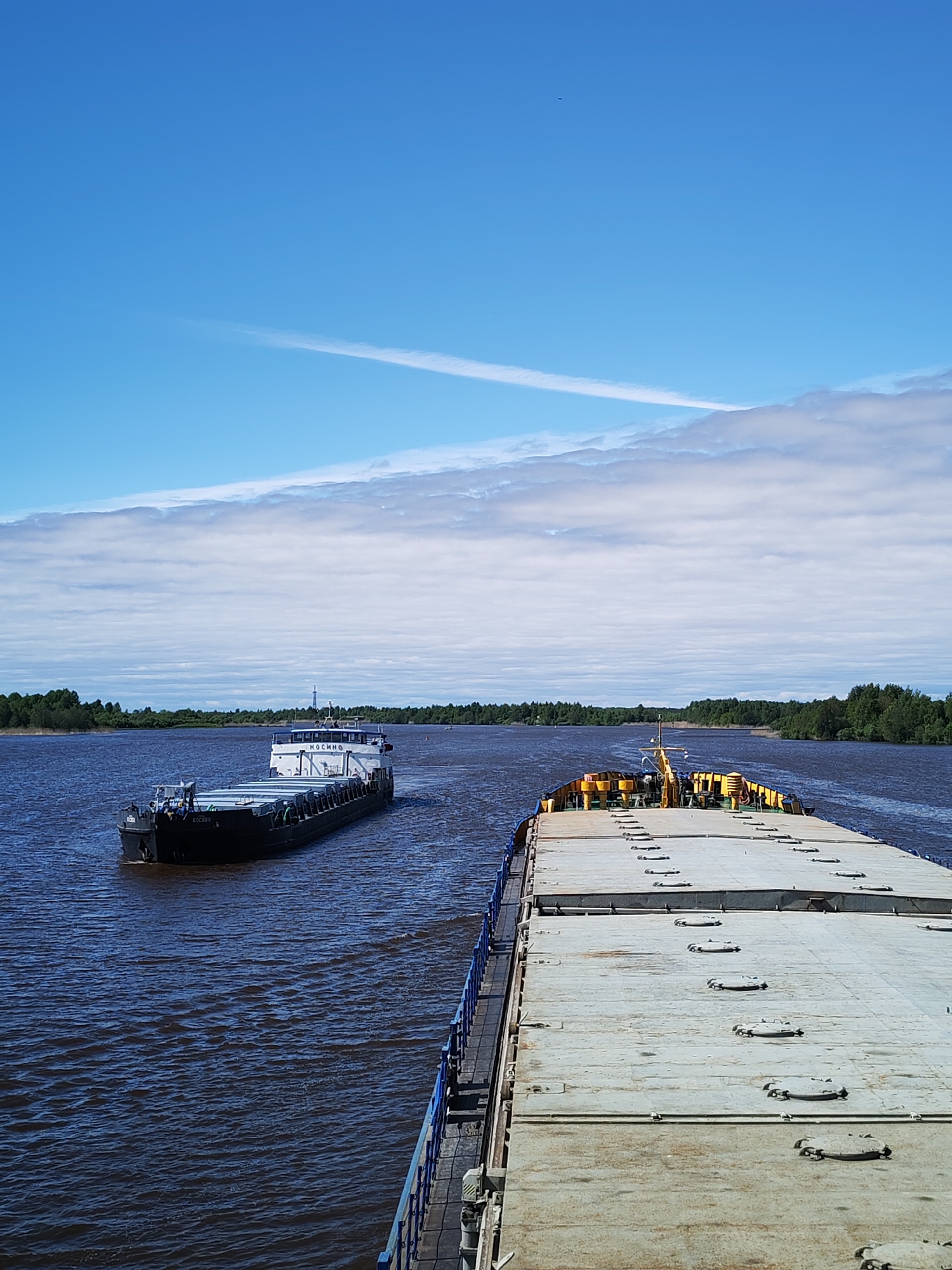 A daytime, medium-shot depicts a river scene near the city of Chaika, Russia. Two barges are visible on the water. One barge is positioned to the left, moving away from the viewer. The other is in the foreground. The latter occupies the bottom half of the frame, showing a gray deck with evenly spaced circular hatches. A blue railing runs along the side of the barge.
The river extends to the horizon, flanked by dense green forests on both sides. Above, a blue sky is partially covered with streaks of clouds and contrails.