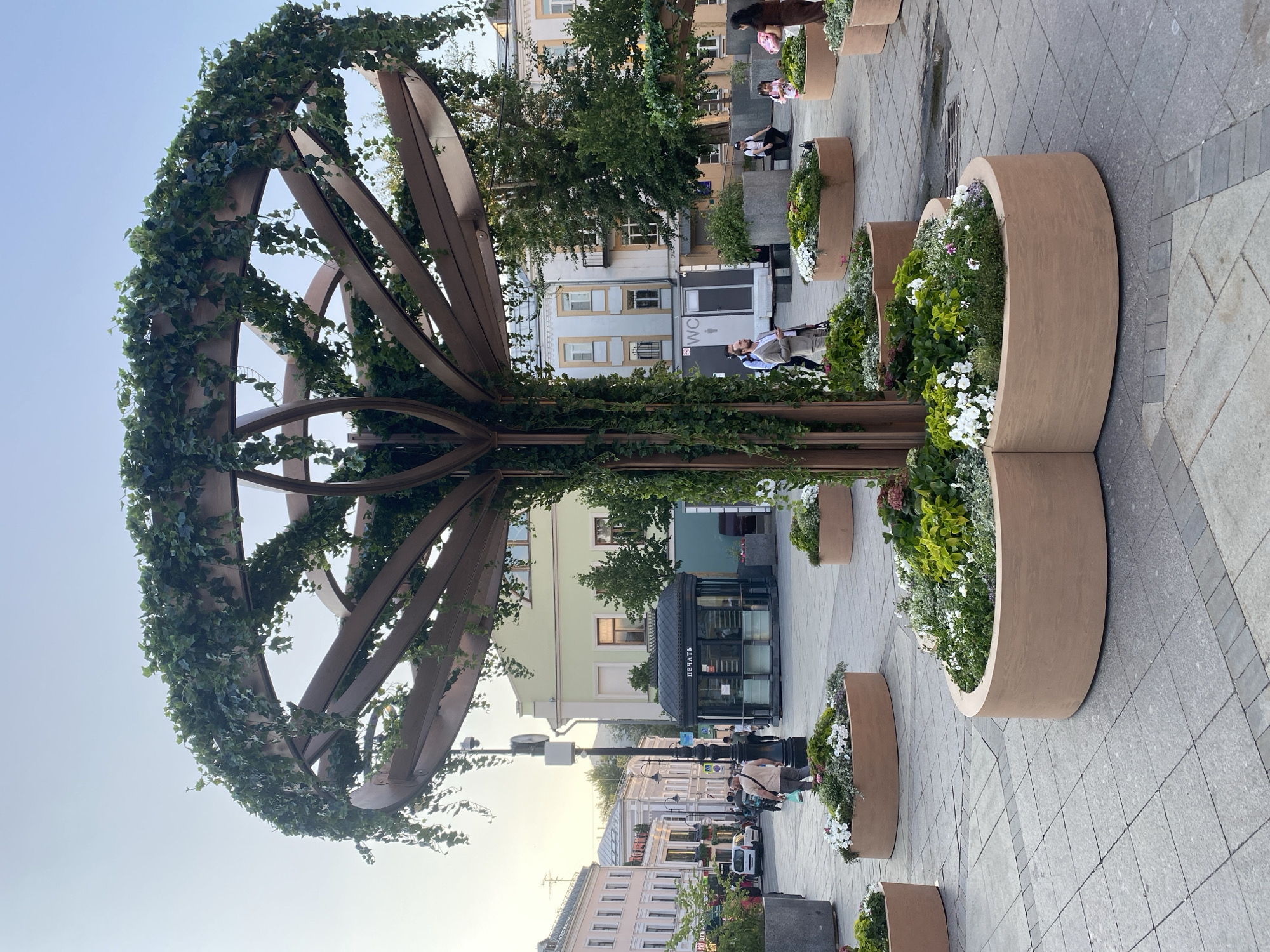 The image depicts a charming outdoor scene, likely a pedestrian street in Moscow, Russia. The foreground is dominated by a large, artistic structure resembling a tree or a globe, crafted from wood and adorned with vibrant green ivy. Surrounding this structure are several large, curved planters filled with an abundance of lush greenery and colorful flowers, including white petunias and other blossoms. The planters are arranged in a decorative pattern along a paved walkway.
In the background, several people are visible, casually strolling and enjoying the outdoor environment. Some are seated near the planters, while others walk along the street. The setting includes buildings with distinct architectural styles, suggesting a well-established urban area. One building features a sign with Cyrillic text, partially obscured, and a kiosk with the word "ПЕЧАТИ" (PECHATI), meaning "PRESS" or "PRINT," is also visible, indicating a newsstand.
The lighting suggests it is either early morning or late afternoon, with a soft, natural light illuminating the scene. The sky is clear and a light blue. The overall atmosphere appears peaceful and relaxed, with people enjoying a pleasant day outdoors in a well-maintained, aesthetically pleasing urban space.