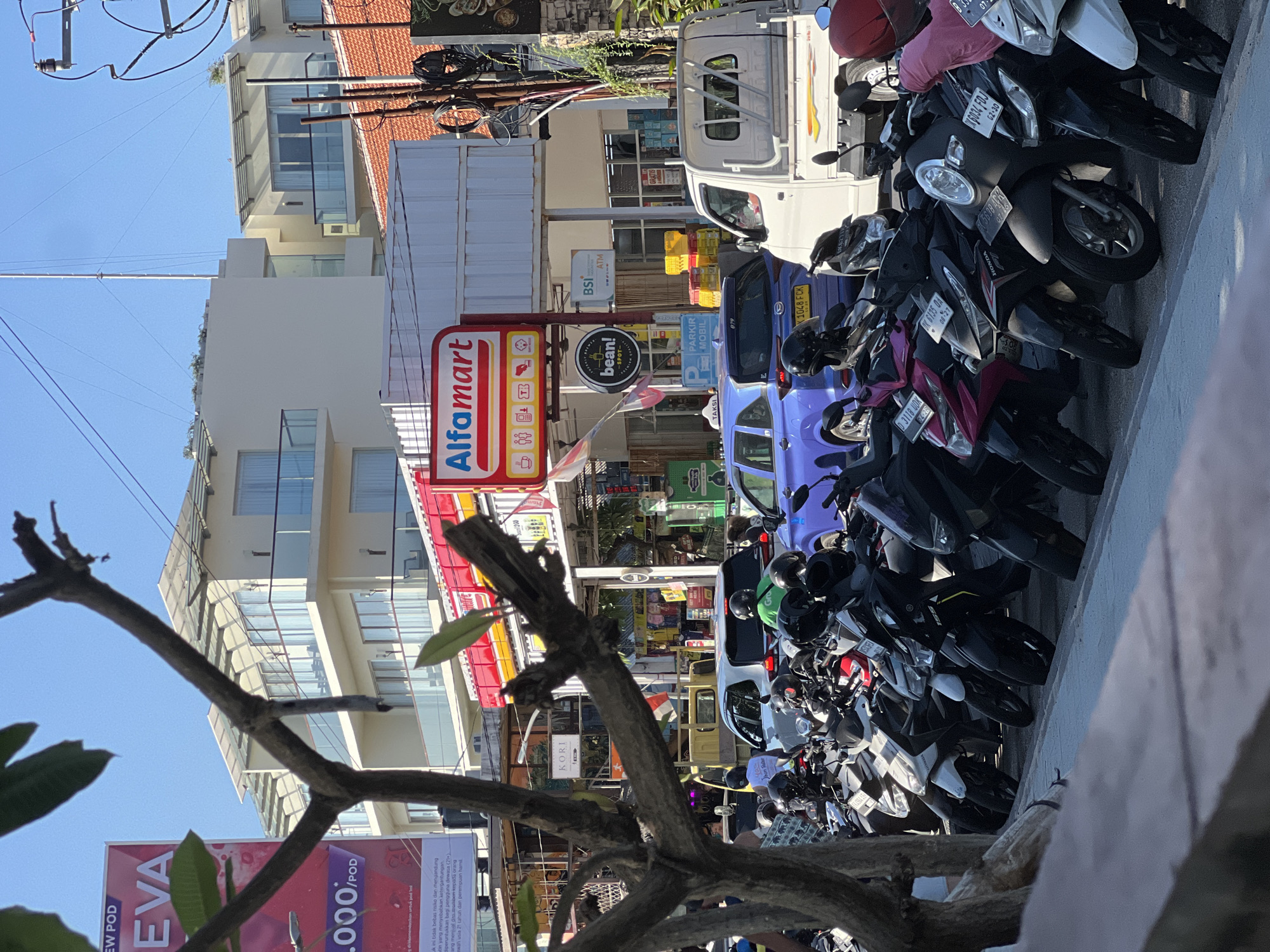 This is a street-level, eye-level, medium shot of a busy commercial area in Kuta, Bali, Indonesia. The scene is dominated by a row of parked motorcycles, with a variety of cars and buildings in the background. The weather appears to be clear and sunny, with a bright blue sky.
In the foreground, a large, dark tree branch with sparse leaves extends across the frame, partially obscuring the view of the street. Below the branch, a line of motorcycles is parked on the side of the road. The motorcycles are of various colors, including black, white, red, and pink. Several riders are visible wearing helmets, and one person is wearing a green shirt.
In the mid-ground, several cars are parked and in motion. A white pickup truck is parked on the right, and a blue sedan is visible to the left. In the background, a white building with multiple stories and balconies stands tall. A red and white Alfamart sign is prominently displayed on the front of a building. Other businesses, including a "bean! SPOT" cafe and a BSI ATM, are also visible. The street is lined with shops and storefronts.
The overall impression is of a vibrant and bustling urban environment, characteristic of a popular tourist destination in Indonesia.