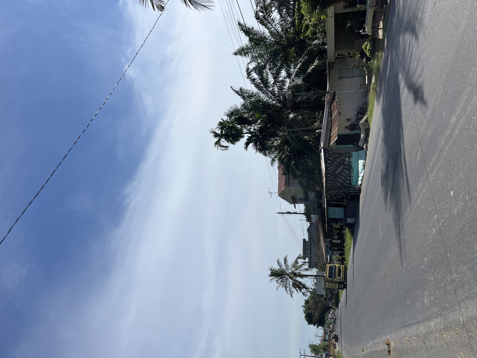 The image depicts a roadside scene in a tropical environment, likely in Bukitrata, Indonesia, under a bright, partially cloudy sky. The perspective is from a low angle, looking upwards and sideways, with the road occupying the right portion of the frame. Along the left side of the road, a row of buildings and lush vegetation, including palm trees, are visible. Some of these structures appear to be simple dwellings or small businesses. Several people are gathered outside one of the buildings, seated or standing. A yellow truck is parked on the side of the road near some of the buildings, and other vehicles are present further down the road, suggesting moderate traffic. The presence of palm trees and the architectural style of the buildings are consistent with a tropical Southeast Asian location. The lighting indicates it is daytime, likely late morning or early afternoon, given the shadows cast. The sky is a vibrant blue with wispy white clouds, suggesting fair weather. A power line runs diagonally across the sky from the upper left corner, a common sight in many populated areas. The overall atmosphere is one of everyday life in a developing rural or suburban area. There is some text visible on a sign above one of the buildings, appearing to be "RAY ARYA".