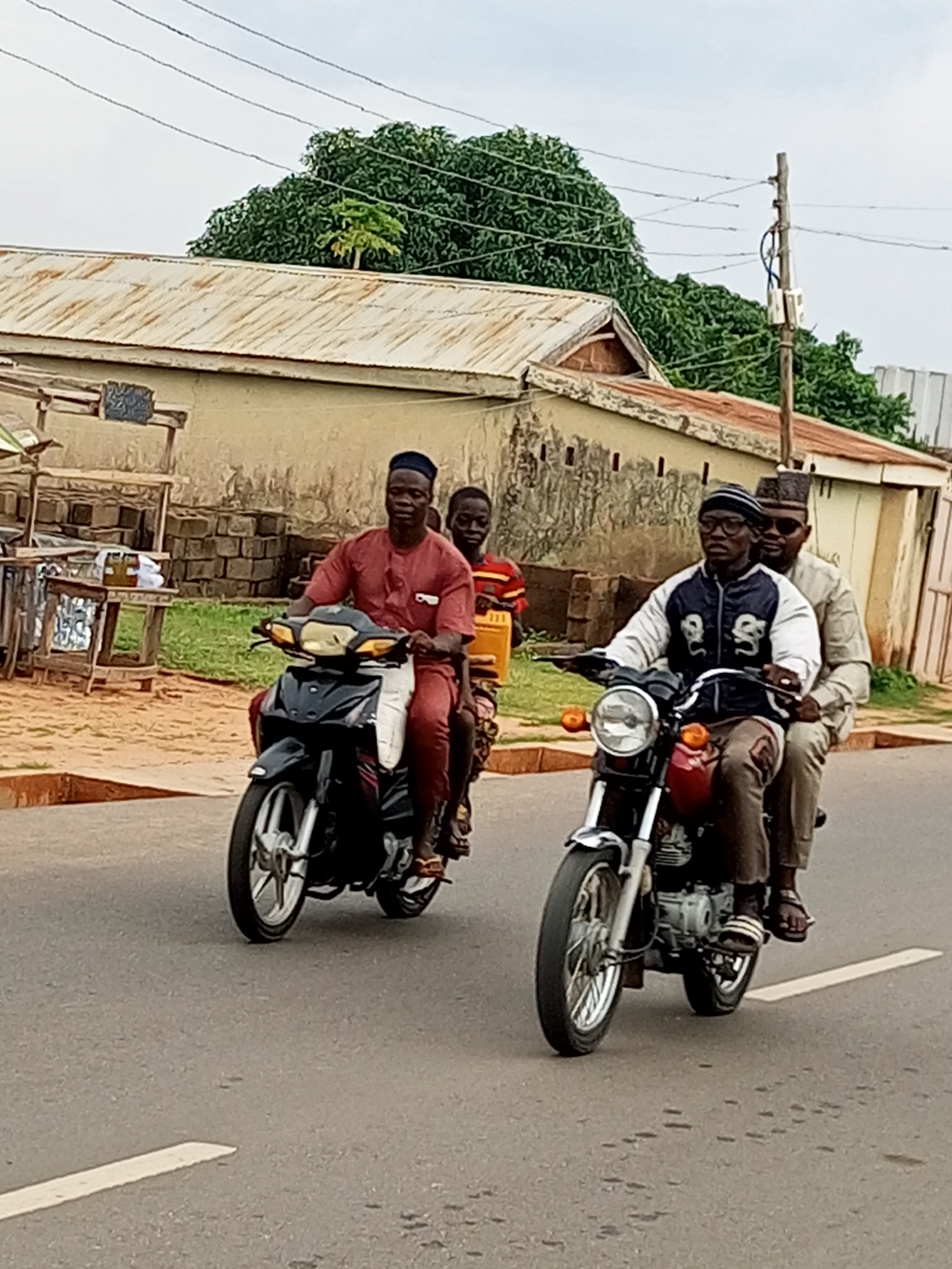Two motorcycles are traveling on a paved road in a rural or semi-urban setting. The motorcycle on the left is black and silver and carries three people: a man in a red shirt and pants driving, a boy in a yellow shirt behind him, and a younger child behind the boy. The motorcycle on the right is red and silver and carries two people: a man in a blue and white jacket and a patterned cap driving, and a man in a light-colored shirt and cap on the back. The environment includes buildings with corrugated metal roofs, trees, and a telephone pole. The sky is overcast.
