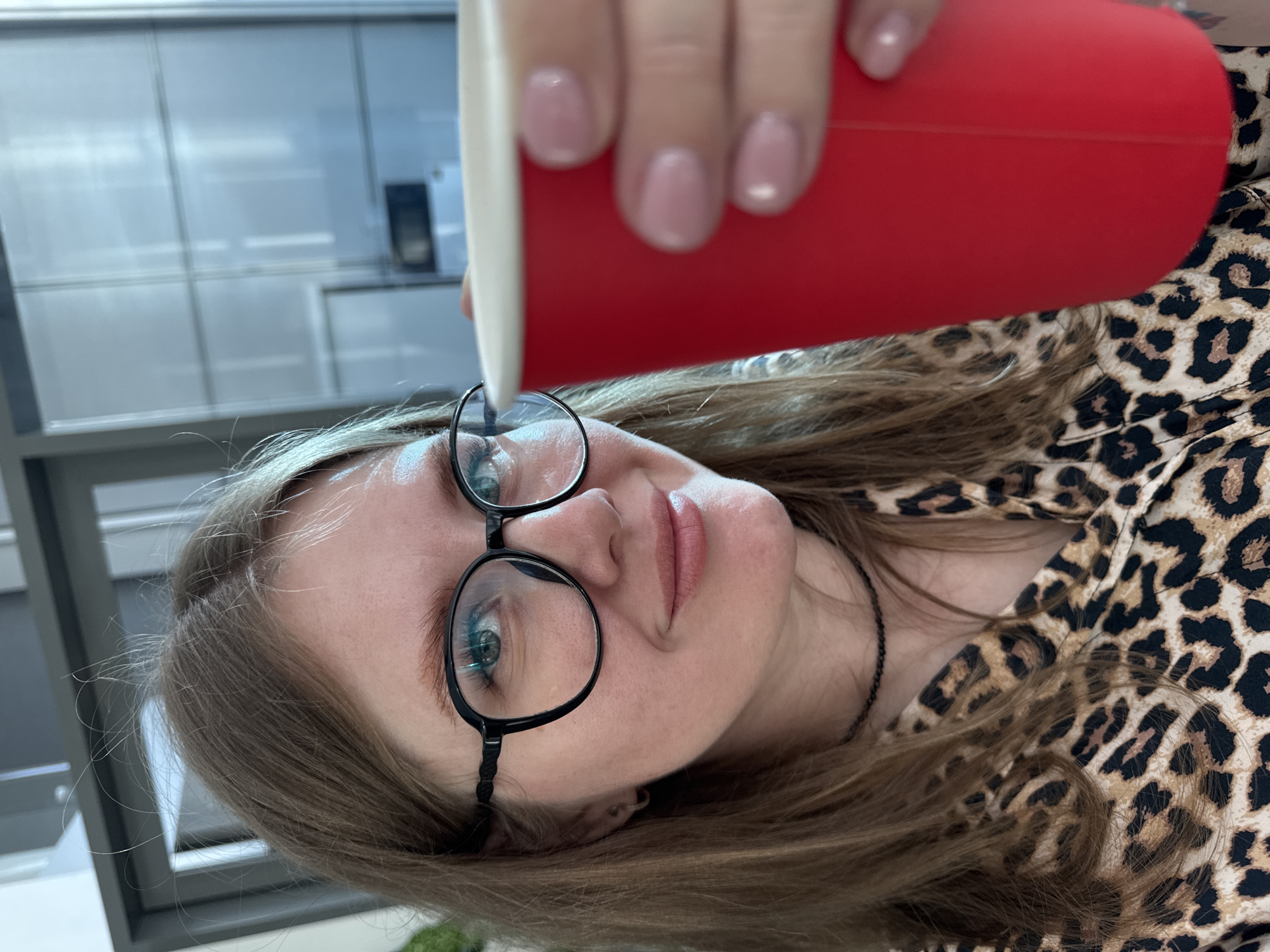 A woman with long blonde hair and wearing round glasses is holding a red cup close to her face. She is wearing a leopard print top. The background shows a modern interior, likely in a building. The location is Perm, Russia.