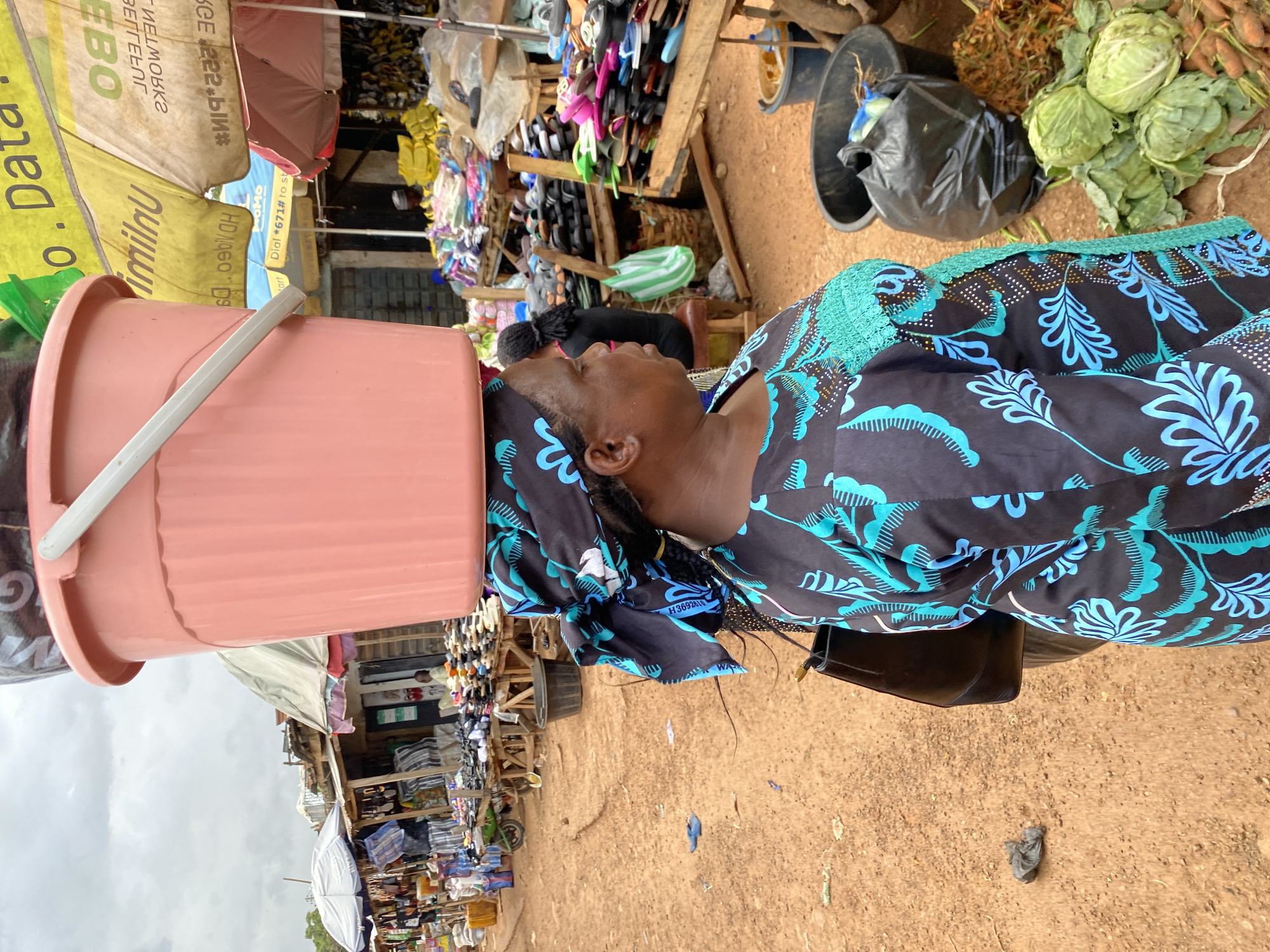 In Bukuru, Nigeria, a woman balances a large pink bucket filled with what appears to be a black plastic bag on her head. She is walking through a bustling outdoor market. The woman is wearing a vibrant black and teal patterned dress and a matching headscarf. She carries a black handbag over her left shoulder. The market stalls display a variety of goods, including footwear, fabric, and fresh produce like cabbage and carrots. The ground is covered in reddish-brown dirt. The sky above is overcast.