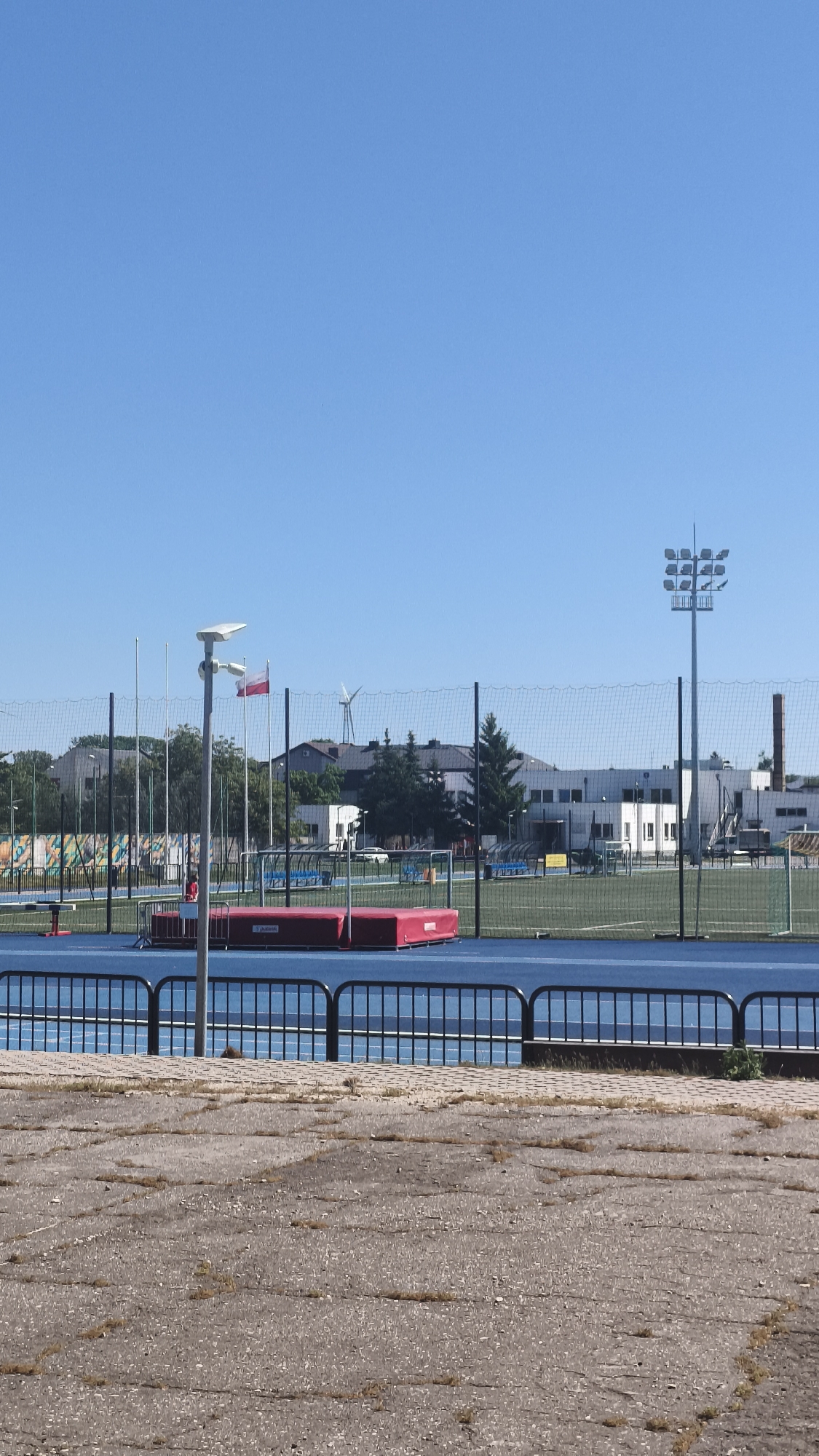 The image shows a view of an athletic field in Nowy Tomyśl, Poland. The foreground features cracked concrete with sparse vegetation. A black metal fence separates the foreground from the track and field area. Behind the fence is a blue track with field event equipment, including a red pole vault landing pad. In the background, netting surrounds a soccer field. Beyond the netting are buildings, trees, a Polish flag, and a wind turbine. A tall light pole stands on the right side of the background. The sky is clear and blue.