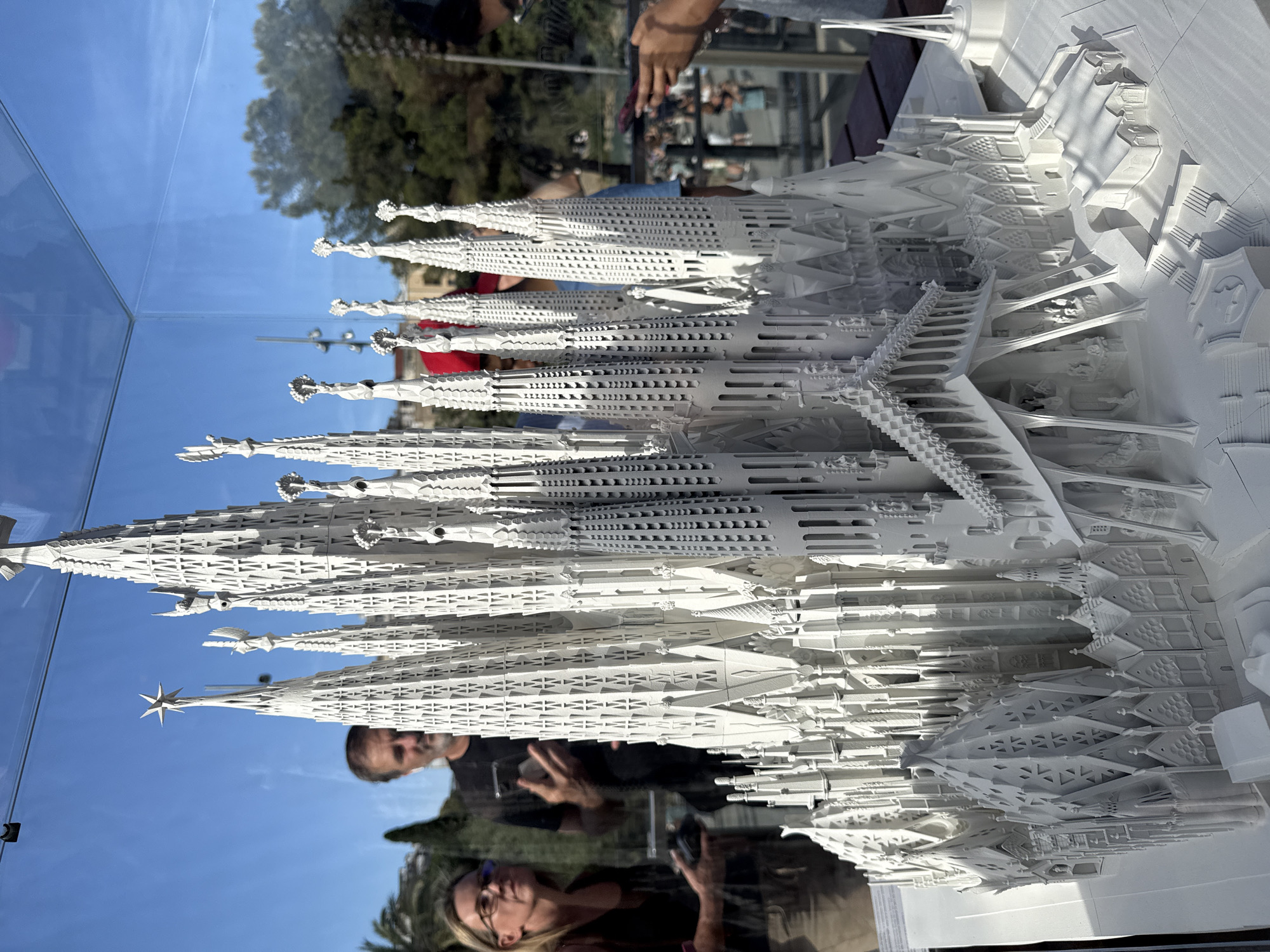 The image shows a detailed white architectural model of the Sagrada Familia in Barcelona, Spain, displayed outdoors under a glass enclosure. The model features intricate spires, facades, and structural elements that mimic the basilica's design. In the background, blurred figures of people are visible, some of whom appear to be looking at the model or taking pictures with their phones. The environment suggests a sunny day, with a clear blue sky and some trees and buildings visible in the distance. The model is placed on a wooden surface, likely a display table.