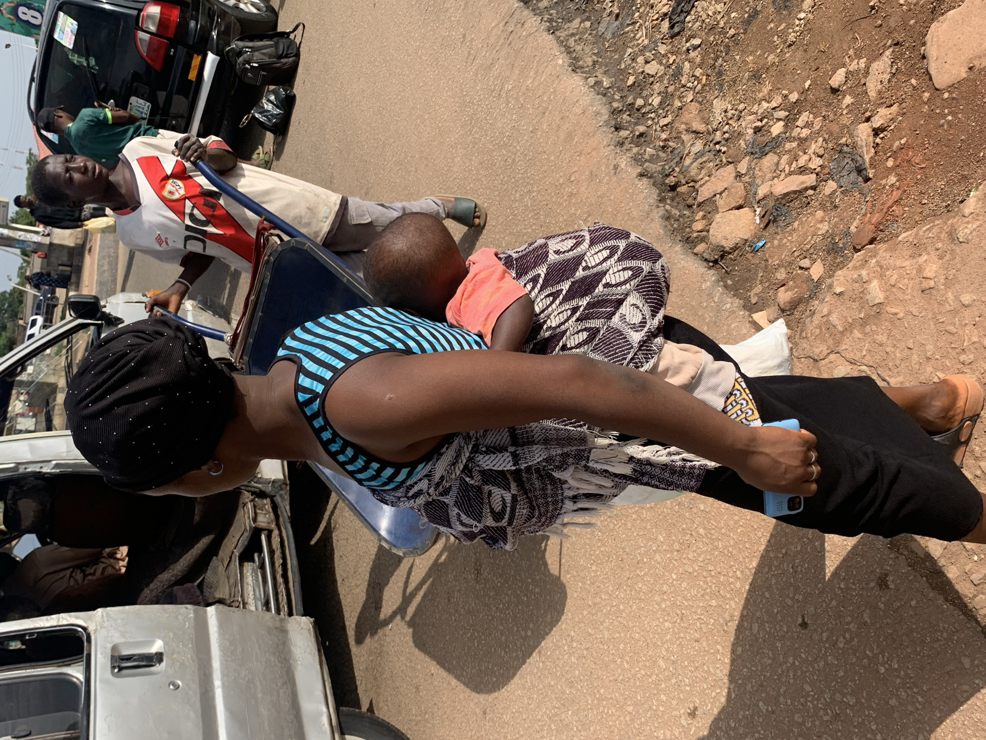 The image depicts a street scene in Gura Top, Nigeria. The primary subjects are three individuals. In the foreground, a person wearing a black headscarf and a black skirt is seated, holding a light blue mobile phone in their right hand. They are wearing a patterned top and a blue and black striped top underneath. Next to them, a child is nestled, wearing an orange shirt and wrapped in a patterned cloth. To the left, behind them, a young man is standing and appears to be leaning on a blue pole. He is wearing a white t-shirt with red accents and grey shorts. In the background, a black SUV is parked, and a person in a green shirt is visible inside.
The scene appears to be taking place during the daytime, with sunlight casting shadows on the ground. The ground is a mix of asphalt and rocky dirt, suggesting an outdoor, possibly unpaved or partially paved, road or roadside. The presence of the SUV and the general setting point towards a busy or semi-busy area. The individuals seem to be engaged in a moment of rest or preparation, perhaps waiting for transport or a break from an activity. The child appears to be asleep or resting. There is no overtly discernible emotion displayed by the adults, but the situation suggests a scene of everyday life in a Nigerian community. The clothing styles and the vehicle are consistent with the location context provided. The image is taken from a slightly elevated perspective, looking down towards the subjects.