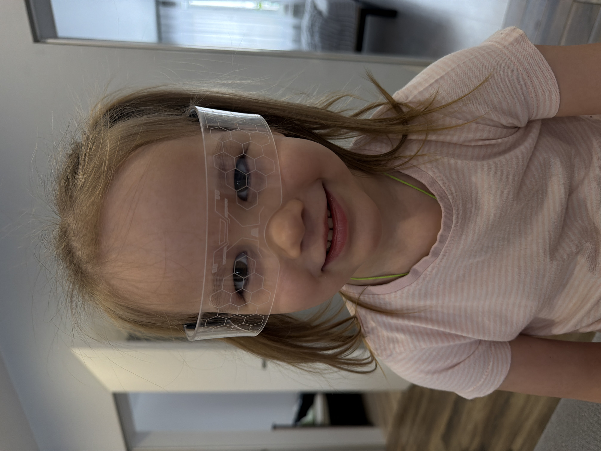 A young girl with light skin and blonde hair smiles while wearing clear, decorative glasses with a honeycomb pattern. She is wearing a light pink and white striped short-sleeved shirt with a neon green collar trim. The background shows a room interior with white walls and what appears to be a wooden floor. There is furniture visible in the background including a bed and a cabinet. No specific details indicate the location is in Chaikovsky, Russia.