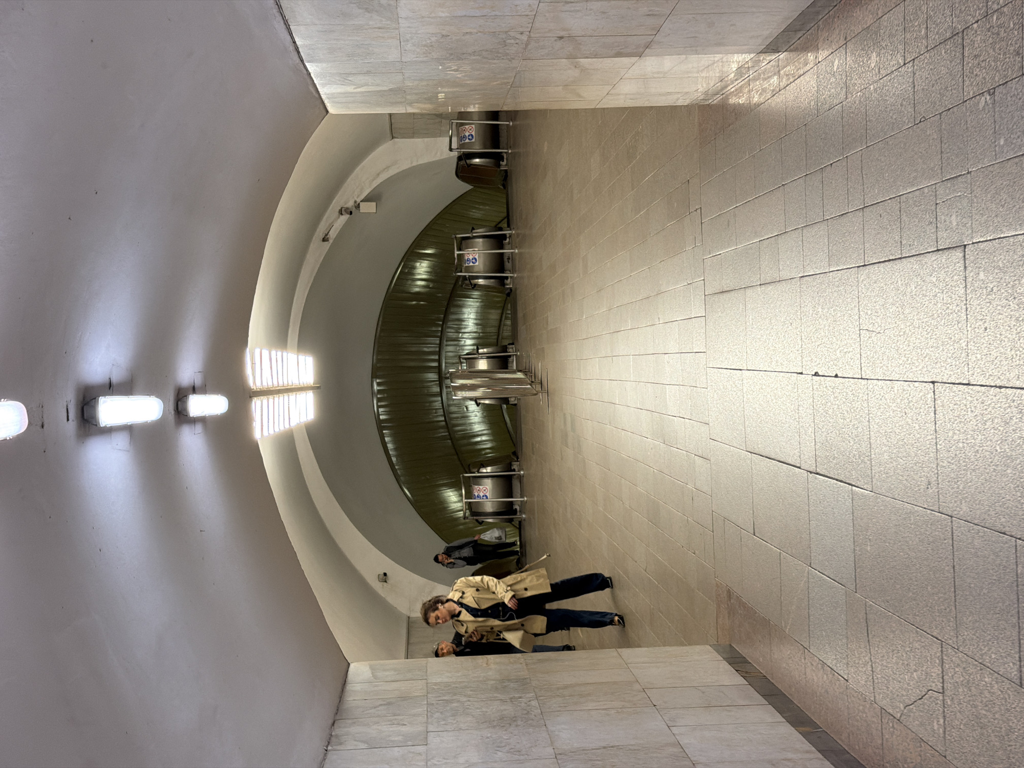 The image captures the interior of a subway station in Moscow, Russia. The walls and floor are lined with light-colored, rectangular stone tiles. The arched ceiling features a series of evenly spaced, rectangular light fixtures and a curved architectural element. A mechanical ventilation system runs overhead. Two individuals are standing on the tiled floor, one wearing a tan trench coat and dark clothing, and the other behind them. Another person is visible in the background.