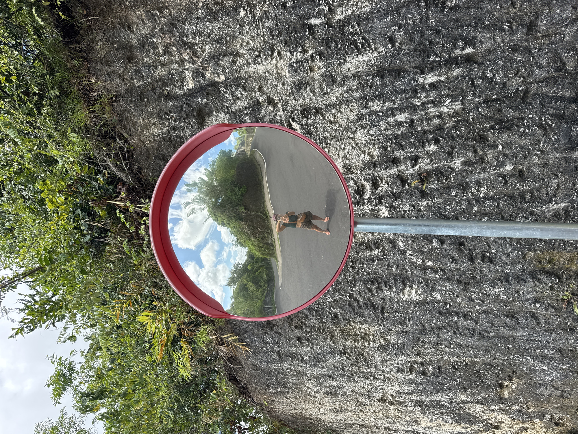 The image shows a convex road mirror mounted on a pole beside a road in Ungasan, Indonesia. The mirror's red frame is visible. The mirror reflects the road, trees, and a cloudy sky. A person is visible in the mirror, standing on the road and taking a picture of themselves with a cell phone. The surrounding environment includes a rough-textured rock wall on one side and dense green vegetation on the other. The image appears to be shot from a slightly angled perspective.