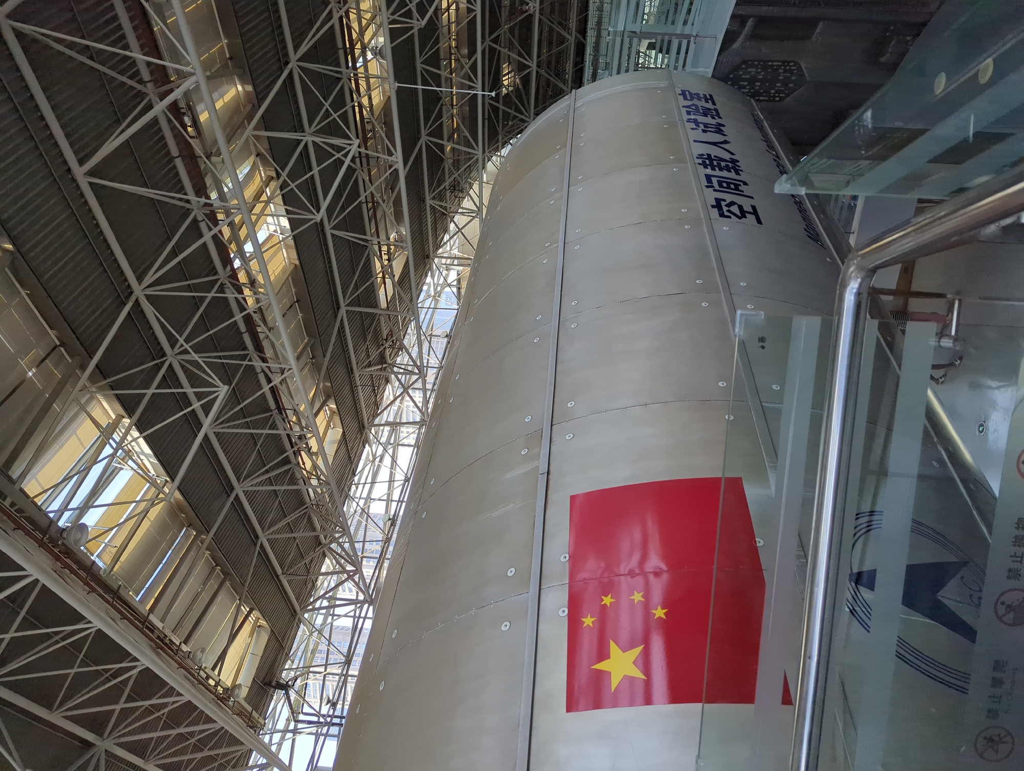 The image shows a large, cylindrical structure, possibly a spacecraft or missile, displayed indoors under a large, industrial-style roof with visible metal trusses and skylights. A portion of the Chinese flag is painted on the side of the cylinder, with five yellow stars on a red background. Below the flag, text in Chinese characters, "空间载人试验室" (Kōngjiān zàirén shìyàn shì), which translates to "Space Manned Test Chamber," is visible. The structure is situated behind a glass barrier with a metal railing, suggesting it is part of a museum or exhibition. The environment appears to be a large hall or hangar. There are no people clearly visible in the immediate foreground or interacting with the object. The lighting is a mix of natural light from the skylights and artificial lighting. The overall impression is a display of a significant technological or space-related artifact. The location is identified as Jiangbeicheng Subdistrict, China.