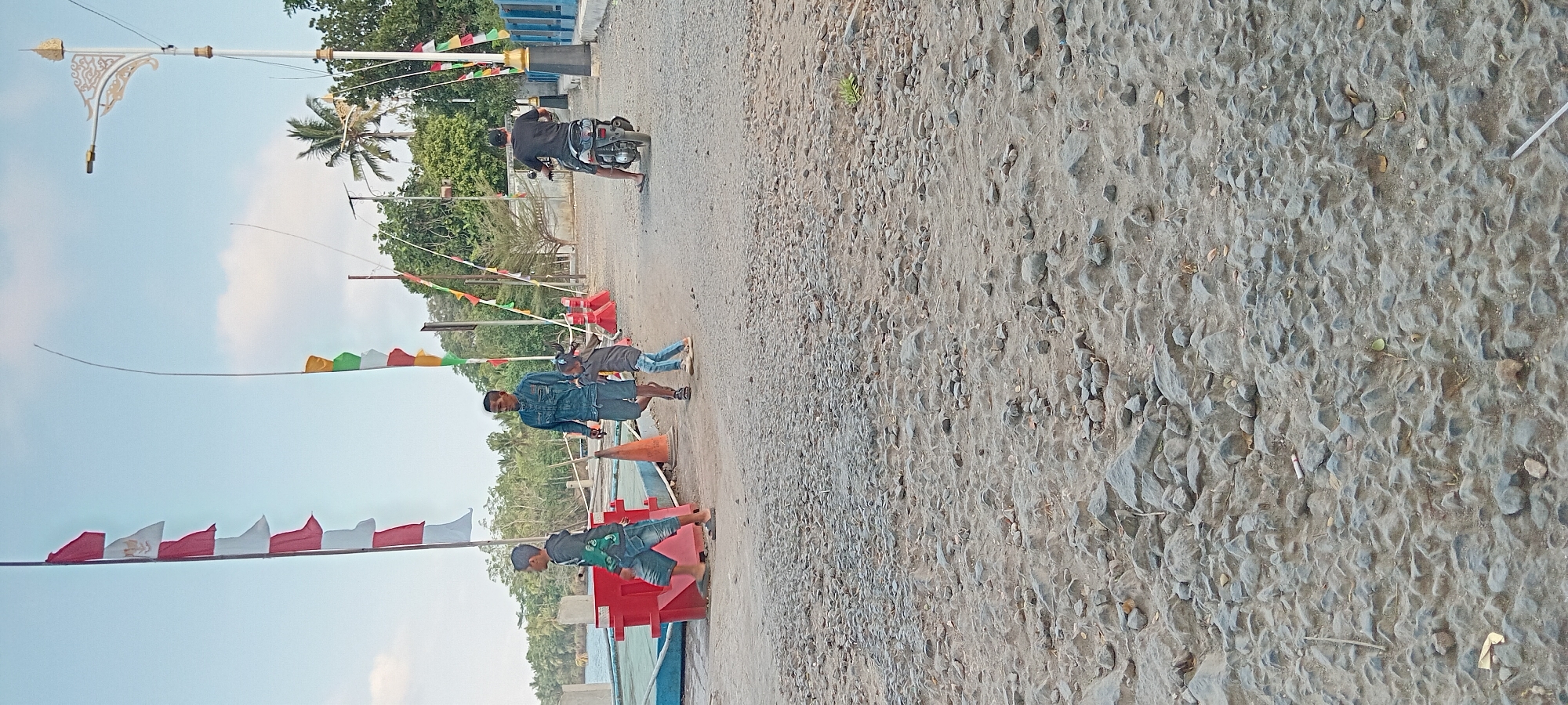 In Kalipucang, Indonesia, a gravel pathway is observed, bordered by a body of water on the left, a red dock structure, and an orange traffic cone. On the pathway, a young male wearing a grey hoodie and denim shorts walks away from the viewer. To his right, a man in a denim jacket and shorts walks towards the viewer, followed closely by a young female in a grey shirt and blue jeans. Further down the path, a person on a motorcycle is traveling away from the viewer. Decorative flags, red and white, are strung on poles along the left side of the pathway, with other colorful flags further along. A lamppost with an ornate design is visible in the background, as are trees and a blue railing. The ground is covered with loose stones and dirt.