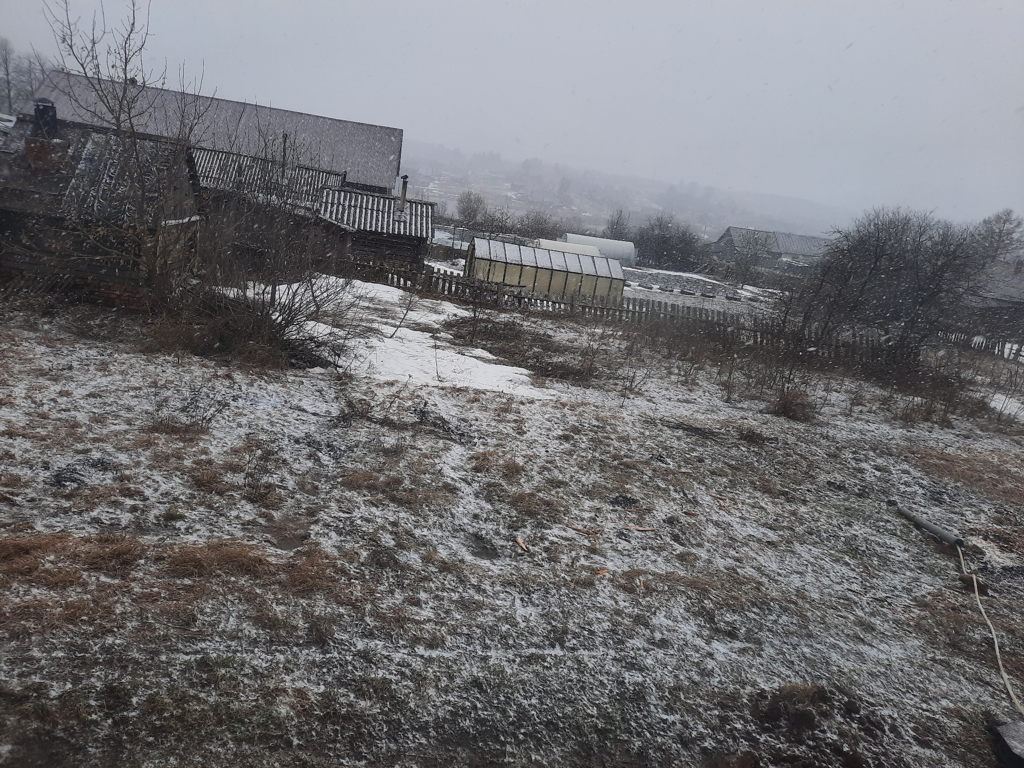 The image depicts a winter scene in Krasnogorskoye, Russia. The landscape is partially covered with snow, showing a mix of snow-covered ground and dry vegetation. Several wooden buildings with corrugated metal roofs are visible in the background. A greenhouse with a clear plastic covering stands behind a wooden fence. Bare trees and shrubs dot the landscape, and snow is falling. A pipe and a white cable are on the right side of the frame in the foreground. The overall atmosphere is overcast and cold.