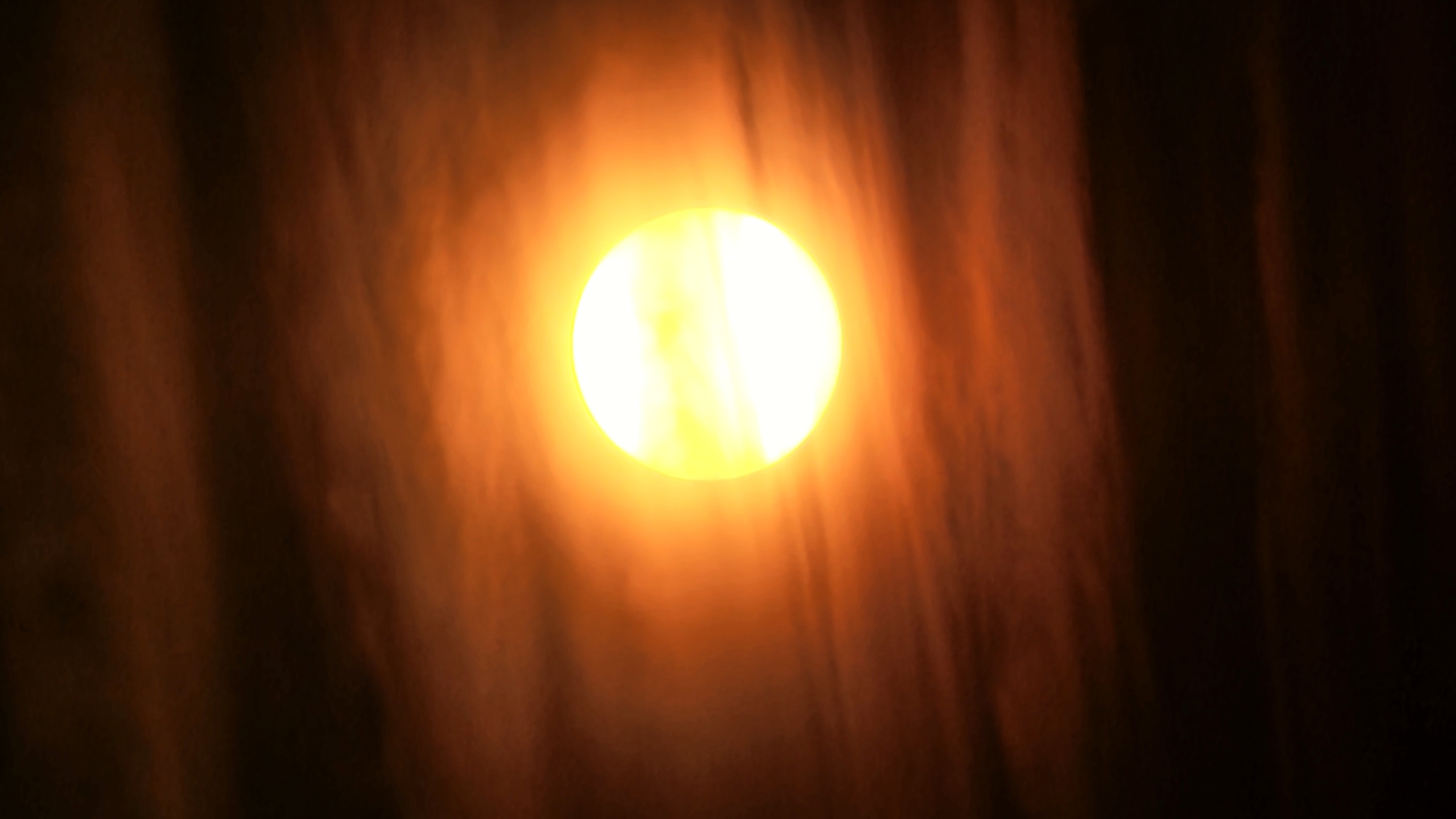A photograph captures a large, bright sun in the center of the frame, partially obscured by wispy clouds. The sun is a brilliant yellow-white orb, radiating a warm glow that bathes the surrounding sky in shades of orange and red. The clouds are dark and textured, appearing as streaks and swirls against the vibrant backdrop of the sun. The overall effect is one of a dramatic sunset or sunrise, with the celestial body dominating the scene. The image does not contain any information about the city of Asan-si, South Korea.