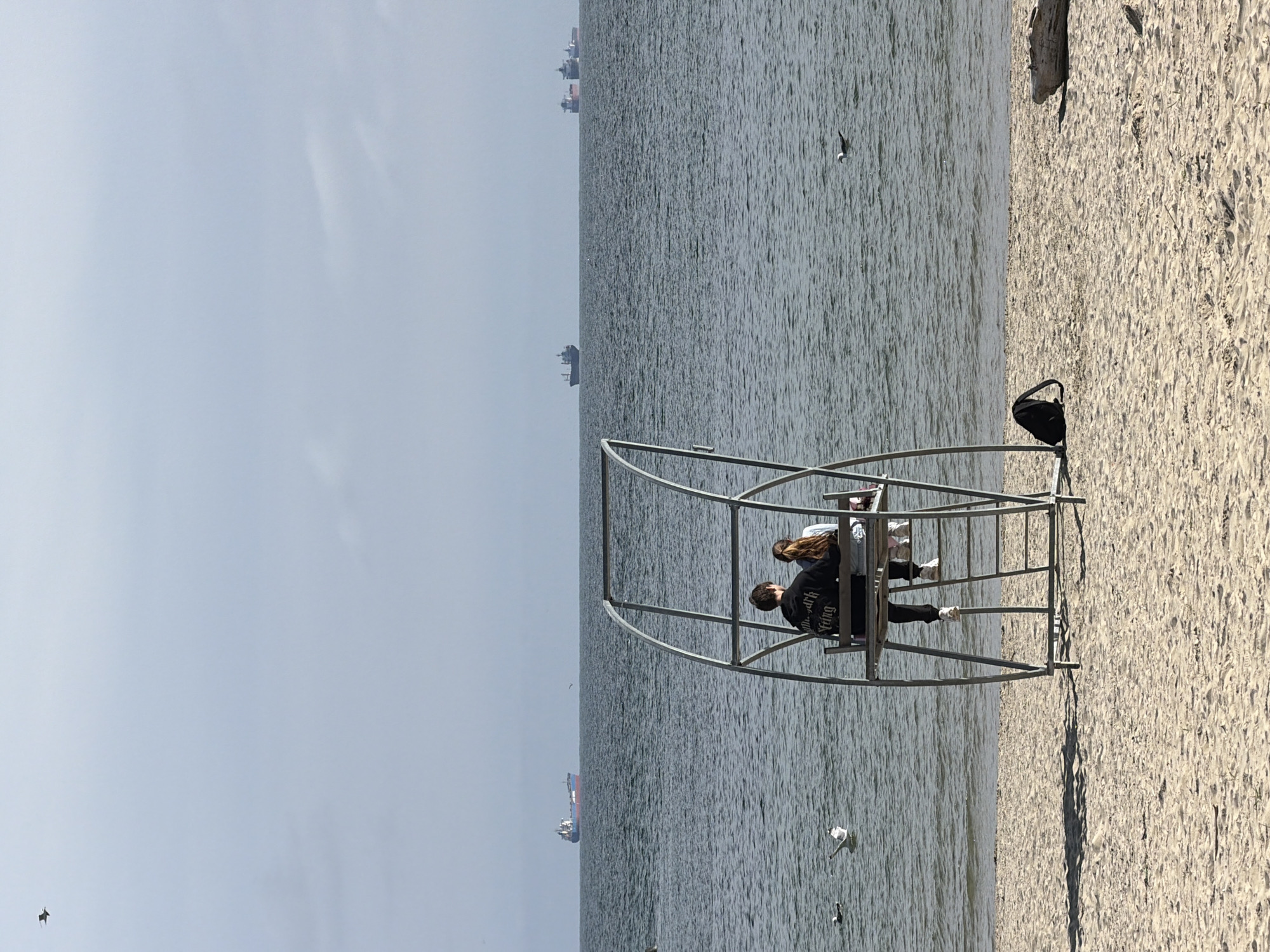 This image captures a serene beach scene in Varna, Bulgaria, on a bright, clear day. The foreground features a sandy beach, where a black backpack rests near a distinctive, modern metallic structure. This open, arched frame, possibly a public art installation or unique seating, extends slightly into the calm, rippling sea.
Two individuals are seated within the metallic structure, facing towards the expanse of water. One person, a man with dark hair, wears a dark sweatshirt with white text visible on the back, which reads "Your New Death is Fling". The other, a woman with lighter hair, is dressed in a light top and dark trousers. They appear to be relaxing and observing the sea.
The midground is dominated by the light gray-blue sea, which stretches to a distant horizon. Several seagulls are visible, with some gracefully floating on the water and one in flight. In the far distance on the horizon, several large cargo ships or tankers are silhouetted against the bright, slightly hazy sky, suggesting a busy shipping lane. The sky itself is a uniform light blue with minimal cloud cover, indicating pleasant, sunny weather. A piece of driftwood is visible on the sand further up the beach. The overall atmosphere is tranquil and contemplative.