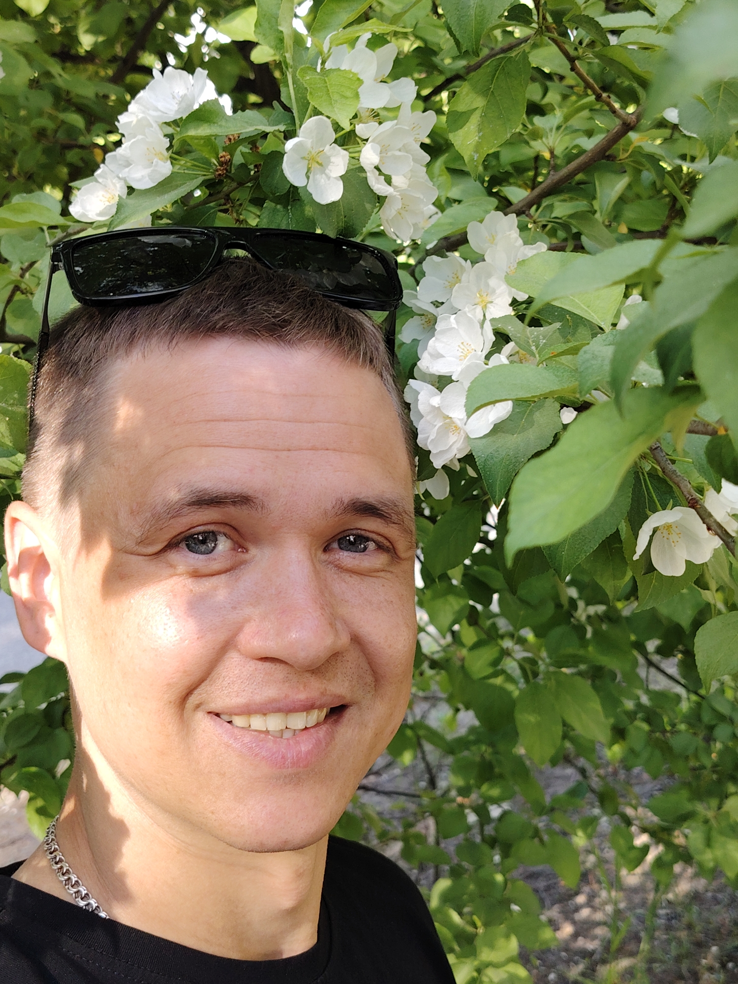 A close-up selfie features a smiling Caucasian male with short, light brown hair and blue eyes, who has sunglasses resting on the top of his head. He is wearing a black t-shirt and a silver chain around his neck. Behind him, lush green foliage and white blossoms fill the background. The photograph was taken outdoors in natural light. The location is in the city of Chaikovsky, Russia.