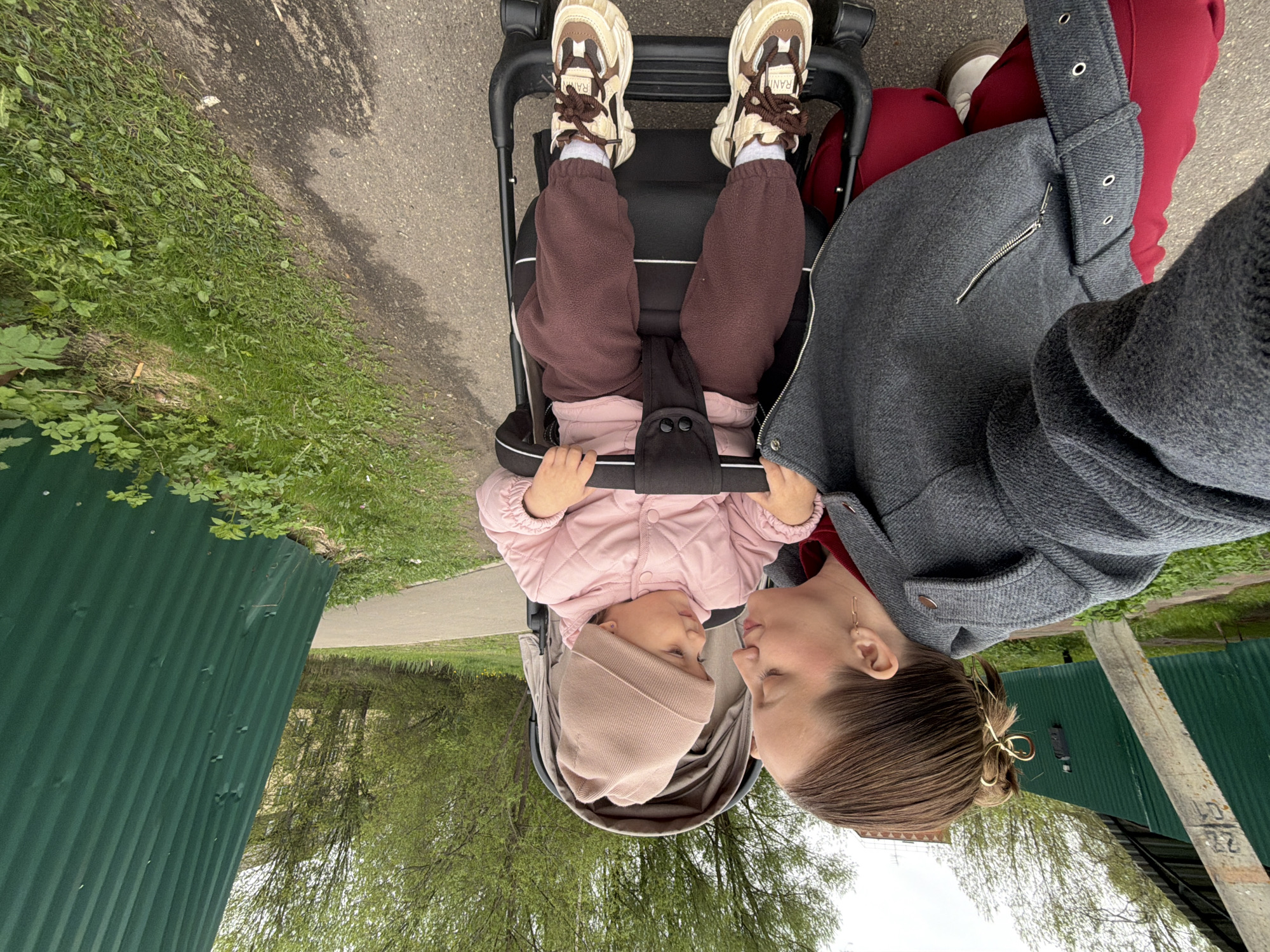 A woman is standing behind a baby in a stroller on a paved path, likely in Reshetnikovo, Russia. The baby is wearing a light pink quilted jacket, maroon pants, tan sneakers, and a light tan beanie. The woman wears a gray coat, and her hair is up with a gold clip. They are facing each other, with their faces close, appearing to interact. The background shows green grass, trees, and a green metal fence. A concrete pole is seen on the right side. The photo is taken from a first-person perspective.