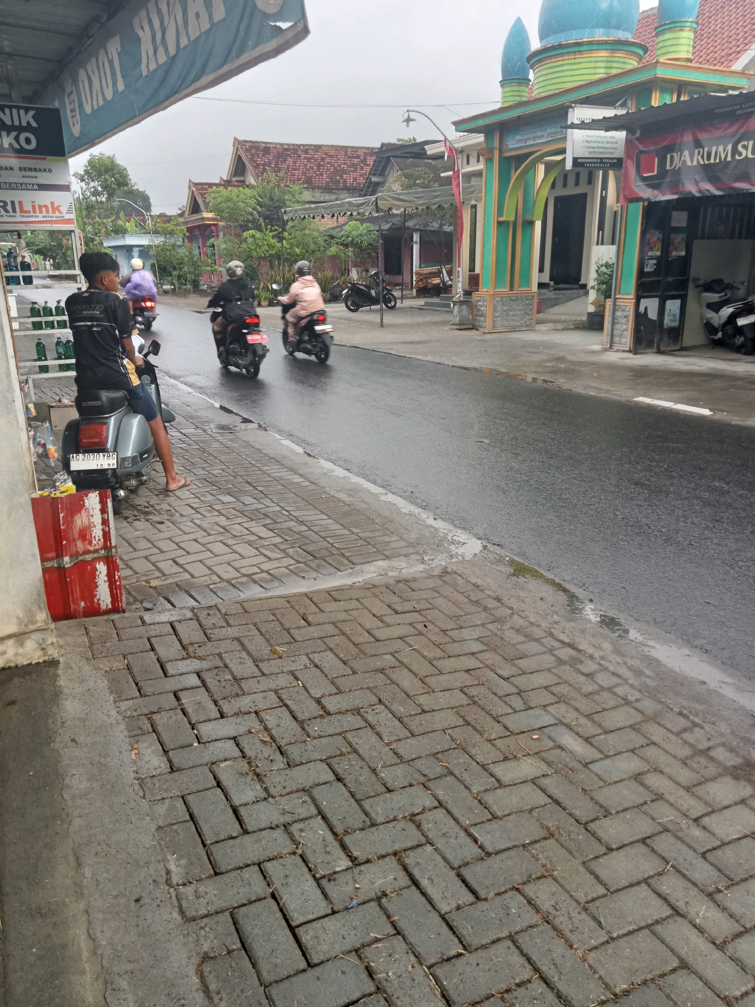 A street scene in Trenggalek, Indonesia, is depicted on an overcast day. In the foreground, a young person sits on a silver scooter parked on a brick sidewalk. The person wears a black t-shirt and blue shorts, with flip-flops on their feet. A red, weathered metal object stands next to the scooter. The sidewalk is laid with interlocking brick pavers and shows signs of moisture, with some leaves and debris scattered on its surface. To the right, the wet asphalt road is visible, with several motorcycles passing by. One motorcycle in the middle ground carries a rider in a black jacket and helmet, and another to the right has a rider in a light-colored jacket and helmet. In the background, a building with green and gold architectural details and blue domes, likely a mosque or religious building, stands prominently. A banner with Indonesian text is displayed on the building's facade. Adjacent to this structure is a storefront with a black sign. Further back, several houses with red-tiled roofs are visible, surrounded by trees and vegetation. The overall atmosphere suggests a typical day in a residential or commercial area of the city.