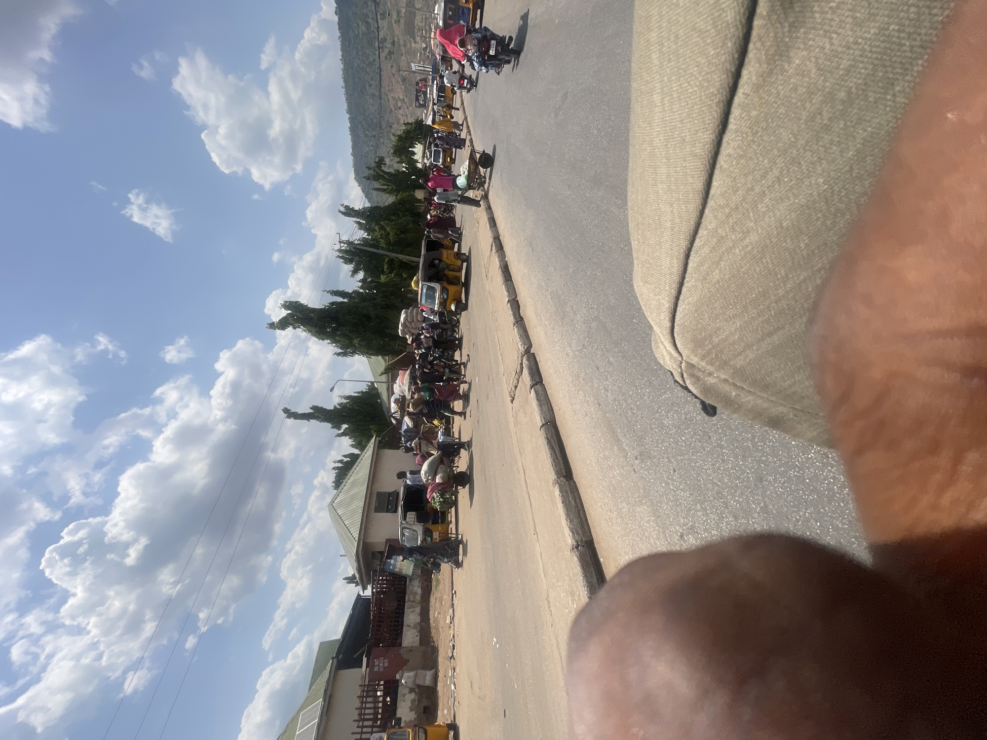 The image, taken from a moving vehicle, presents a bustling street scene in Lokoja, Nigeria. The street is crowded with pedestrians, motorcycles (okadas), and auto-rickshaws (keke napeps) which are common forms of transport. Many pedestrians are carrying goods on their heads, a common practice in the region. Buildings are visible along the road with diverse architectural styles. The sky is blue with scattered clouds. A large mass obscures much of the view from what may be a part of the photographer.