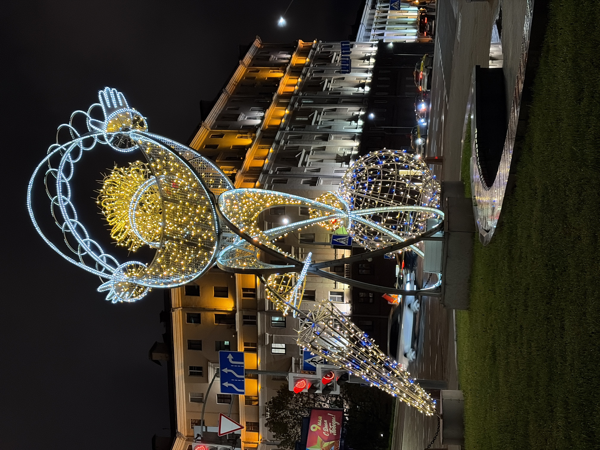 The image shows a nighttime scene in an urban environment. An ornate, lighted sculpture dominates the foreground, consisting of interconnected wireframe structures adorned with white and yellow lights. In the background, a large, multi-story building with illuminated windows stands. Traffic signs are visible, and the street appears wet. A patch of green, possibly grass or artificial turf, is in the lower right corner. The scene suggests a city street decorated for an event or holiday. Location is Unknown, Unknown.