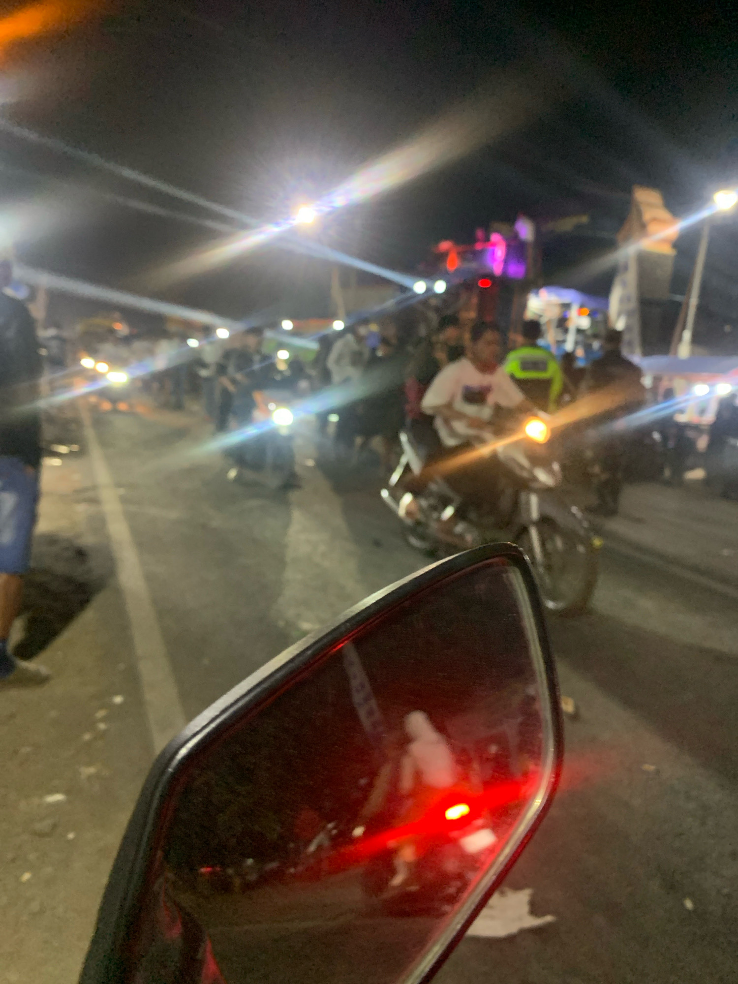 In Talangsuko, Indonesia, at night, a blurry scene depicts a street filled with people and motorcycles. The foreground is dominated by a motorcycle's side mirror, reflecting a red taillight and a ghostly image of a person. Beyond the mirror, the street is illuminated by numerous bright lights, including streetlights and motorcycle headlights, creating lens flares. A white line marks the edge of the road. A crowd of people are visible, some standing and others on motorcycles. One person in the mid-ground is wearing a white shirt and another in the background is wearing a bright green vest. In the distance, there are structures with colorful lights, possibly stalls or stage lighting for an event. The ground appears wet, reflecting the surrounding lights.