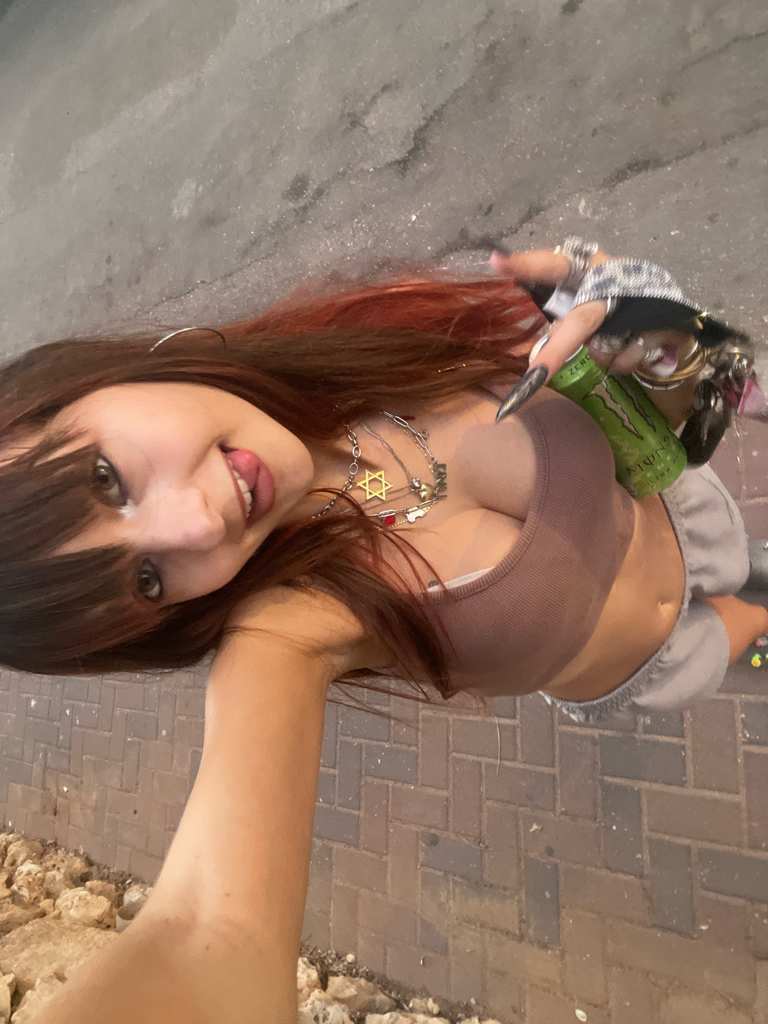 The image shows a young woman taking a selfie outdoors. She has long, brown hair with red highlights and bangs. She is wearing a brown ribbed crop top, grey sweatpants, and multiple necklaces, including one with a Star of David pendant. She is also holding a green can of Monster Ultra energy drink. Her nails are long and pointed. The background appears to be a paved walkway with some rocks and debris. The lighting suggests it is daytime. The woman is smiling and sticking her tongue out, indicating a playful or casual mood. The setting seems to be a street or public area. Visible text on the can reads "MONSTER ULTRA ZERO." The location context is Raanana, Israel.