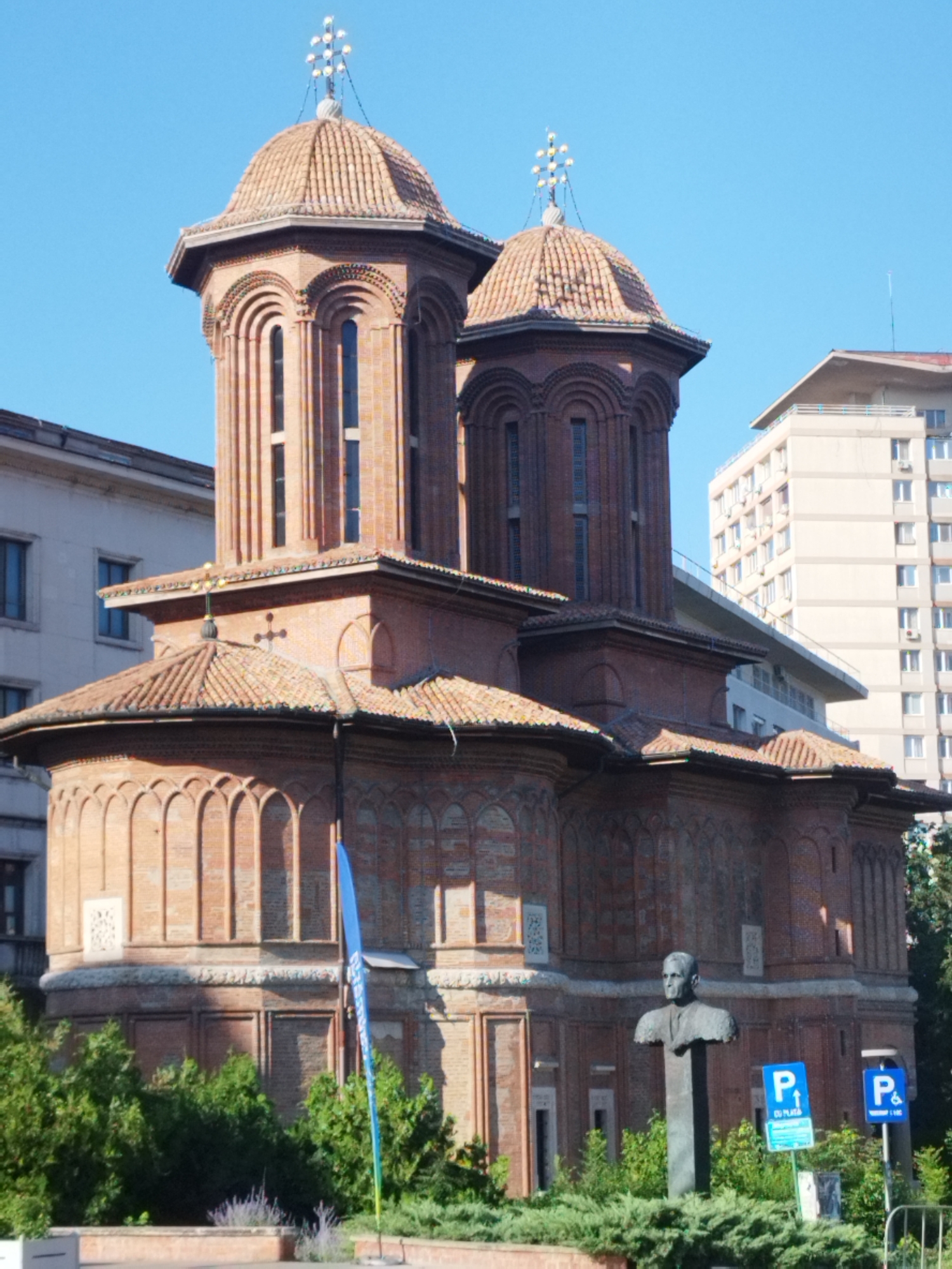 The image depicts the Stavropoleos Monastery Church, a religious building in Bucharest, Romania, situated in the Old Town. The church is characterized by its distinctive red brick architecture, adorned with carved details and arched windows. It features multiple domes, with the central dome topped by a cross. In the foreground, a bust of a man stands on a pedestal, surrounded by lush greenery and bushes. Two blue parking signs are visible, one indicating parking for a disabled person. The sky above is clear and blue, suggesting a sunny day. In the background, modern buildings stand in contrast to the historical church, indicating an urban environment. No people are actively engaged in any discernible actions. The scene is static and appears to be a photograph taken during daylight hours.