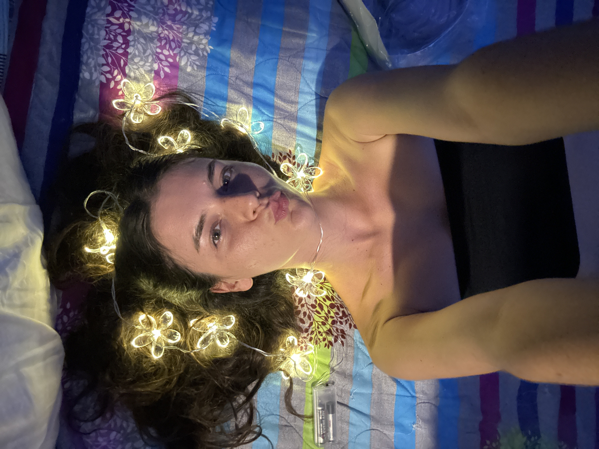 A young woman with long, dark hair is lying on her back on a striped bedspread. She is wearing a black strapless top and has flower-shaped lights woven into her hair. Her head is tilted back, and she is puckering her lips as if for a kiss, looking directly at the camera. A battery pack for the lights is visible to her left. The scene appears to be indoors, possibly in a bedroom, and there is no indication of the city of Mũi Né, Vietnam.