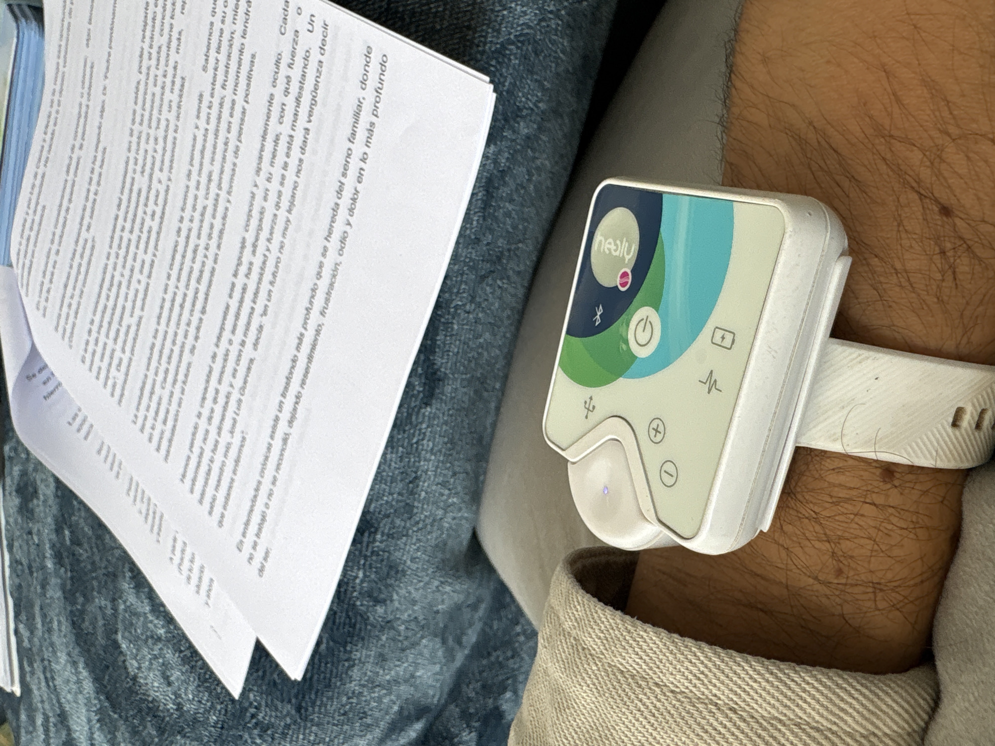 The image shows a person's wrist with a white Healy device attached, positioned next to a stack of papers with Spanish text. The papers contain text on subjects like emotions, diseases and family ties. The Healy device displays a logo and other symbols such as Bluetooth, power and battery symbols. The person is wearing a light-colored garment. The location is undetermined, and there is no information to indicate it is in Puebla, Mexico.
