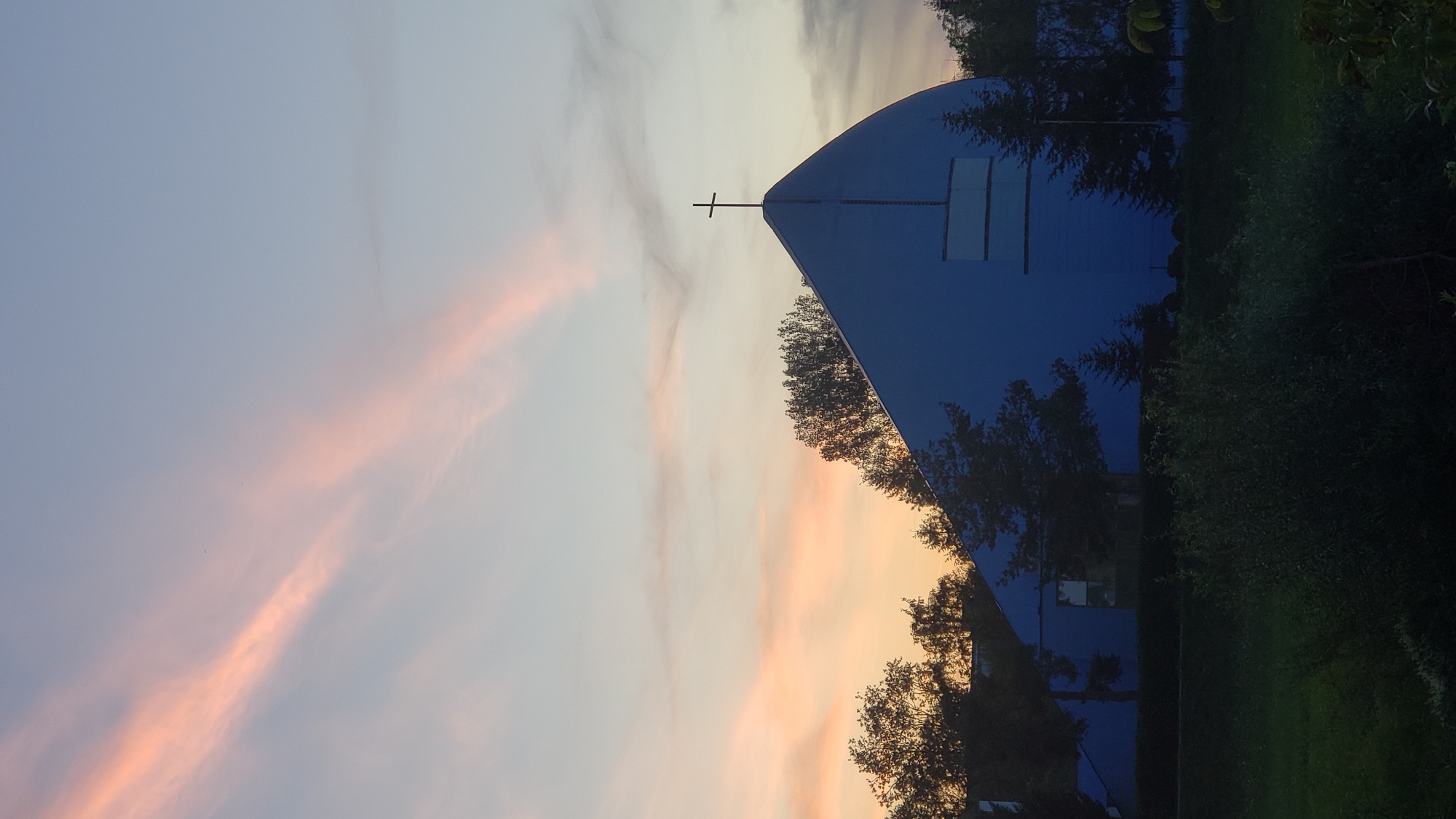 The image shows a modern, blue church with a distinctive curved roof, topped by a simple cross. The church is situated amidst trees, with a manicured lawn and dense bushes in the foreground. The sky displays a soft gradient of colors, transitioning from a pale blue at the top to warm oranges and pinks near the horizon, suggesting either sunrise or sunset. The overall scene is serene and architectural, with the natural elements of trees and sky complementing the man-made structure. This church is identifiable as the Church of the Holy Spirit in Prešov, Slovakia, a notable example of modern religious architecture in the city.