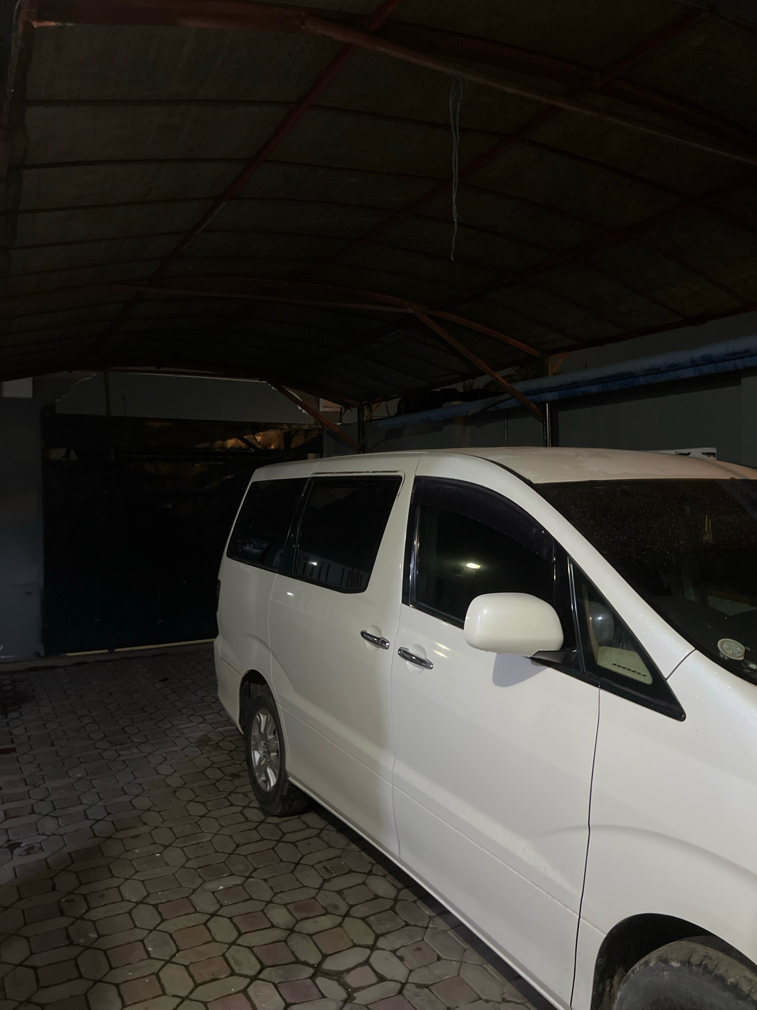 A white minivan is parked on a paver driveway under a covered structure. The structure appears to be an open-air garage with a corrugated metal roof supported by wooden beams. The walls of the structure are made of concrete blocks. To the left of the minivan is a large, dark, metal garage door. The driveway is wet, suggesting recent rain. The scene appears to be in an urban or suburban area of Dar es Salaam, Tanzania.