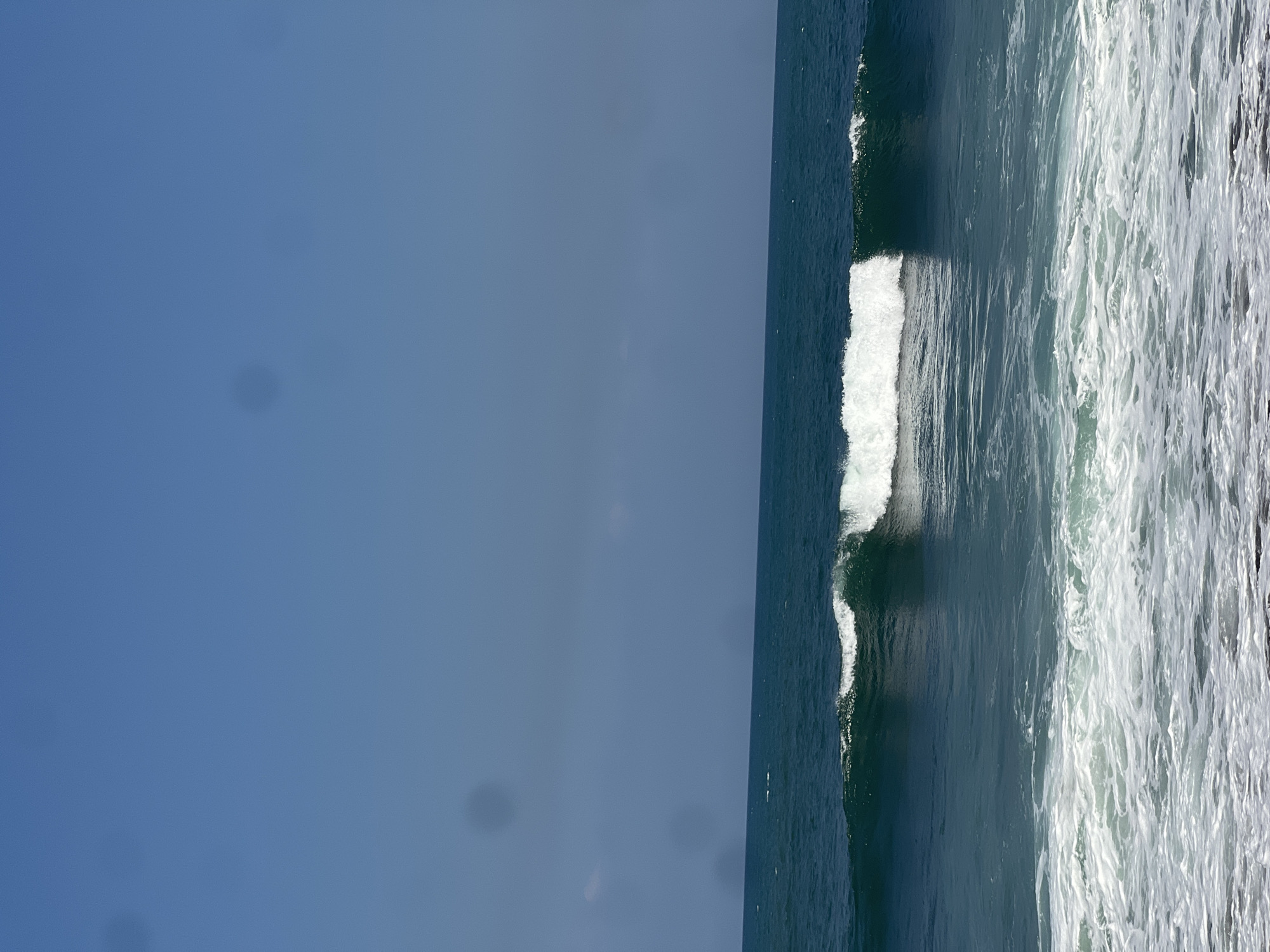 A serene ocean scene unfolds under a clear blue sky. The water is a deep teal, with a gentle wave breaking in the mid-ground, its crest capped with white foam. Closer to the foreground, the water churns into a frothy expanse of white surf. The horizon line is sharp and distinct, separating the vast ocean from the expansive sky. The image appears to be taken from a slightly elevated perspective, possibly a beach or cliff overlooking the water. The location is identified as Mengwi, Indonesia, suggesting a tropical or subtropical coastal environment. There are no people or specific man-made objects visible in the frame, focusing entirely on the natural beauty of the ocean and sky. There are a few dark circular spots in the sky, likely due to a lens issue or something on the camera.