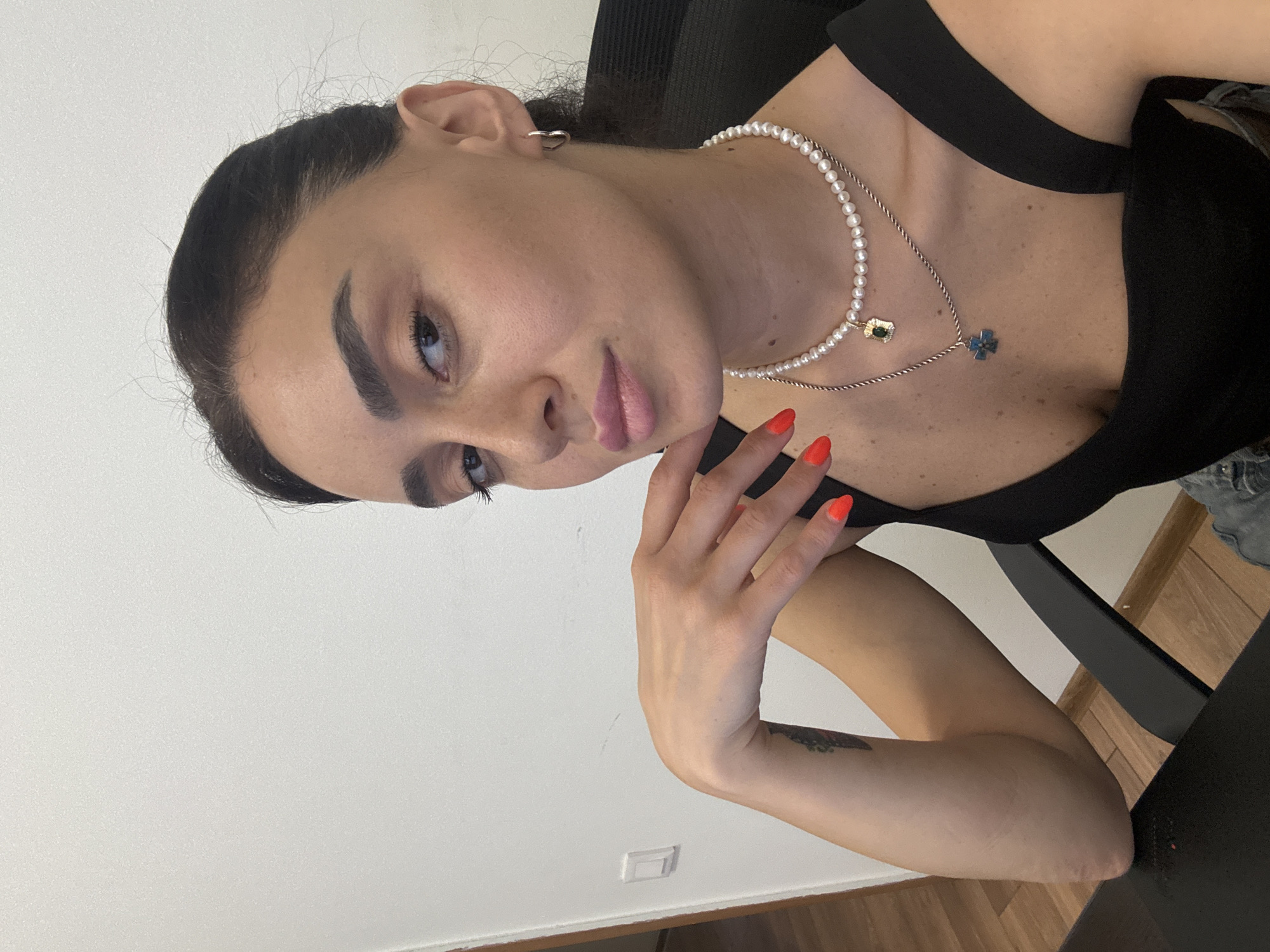 A young woman with dark hair pulled back is seated at a desk, looking at the camera. She is wearing a black top with two necklaces, one with pearls and a small pendant, and another with a blue cross pendant. Her nails are painted bright orange. Her left hand rests on her chin, and her arm is bent at the elbow. A tattoo is visible on her left forearm. The background is a plain white wall with a light switch. A black office chair and a dark desk are also visible. The scene appears to be indoors, possibly in an office or apartment. There is no indication of the location being in the city of Tbilisi, Georgia.