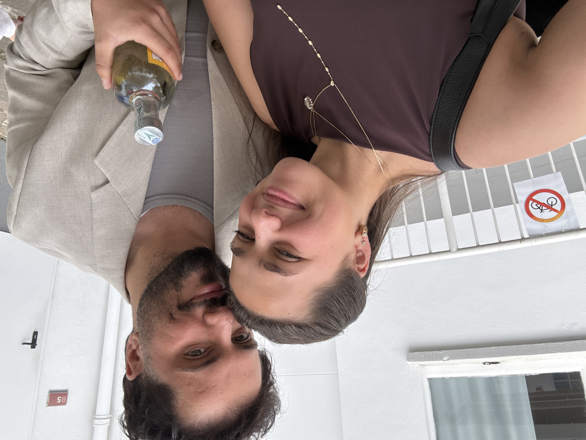 A man and a woman are captured in a close-up, horizontal portrait, smiling gently at the camera. The image appears to be rotated, showing the subjects with their heads towards the left side of the frame. The man, with dark hair and a beard, is wearing a light-colored blazer over a ribbed grey top. He holds a green-tinted glass bottle with a white and blue cap in his right hand. The woman, with dark hair pulled back, is wearing a dark brown sleeveless top, complemented by a delicate gold necklace with a central pendant and another longer gold chain. She also wears small gold earrings. A black shoulder bag strap is visible over her right shoulder.
They are positioned outdoors against a plain white building facade. In the background, to the man's left, a small black fixture is mounted on the wall. A red sign with "B5" in white text is also visible on the wall. To the woman's right, a white metal railing or grate is present, and next to it, a white circular sign with a red border and a diagonal line through a bicycle icon clearly indicates "no bicycles" or "bicycle prohibited." Below the railing, a window with curtains inside can be seen.
The lighting is bright and even, suggesting a daytime shot. Both individuals appear relaxed and happy. The setting, against a white urban wall with utilitarian signs and railings, combined with the "no bicycle" sign, suggests a casual street or building exterior in a city like Milan, Italy.