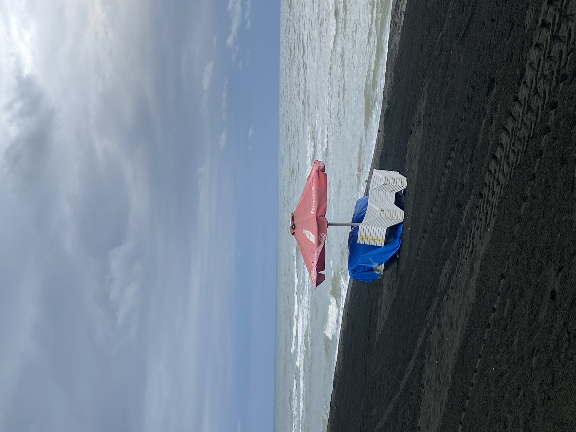 The image depicts a deserted black sand beach in Ureki, Georgia, under a cloudy sky. The sea is calm with moderate waves. In the foreground, a stack of white lounge chairs is covered with a blue tarp, partially hidden by a red umbrella with a Coca-Cola logo. Tire tracks are visible in the dark sand, suggesting recent vehicle activity. The scene conveys a sense of quietude and anticipation, as if awaiting the arrival of beachgoers.