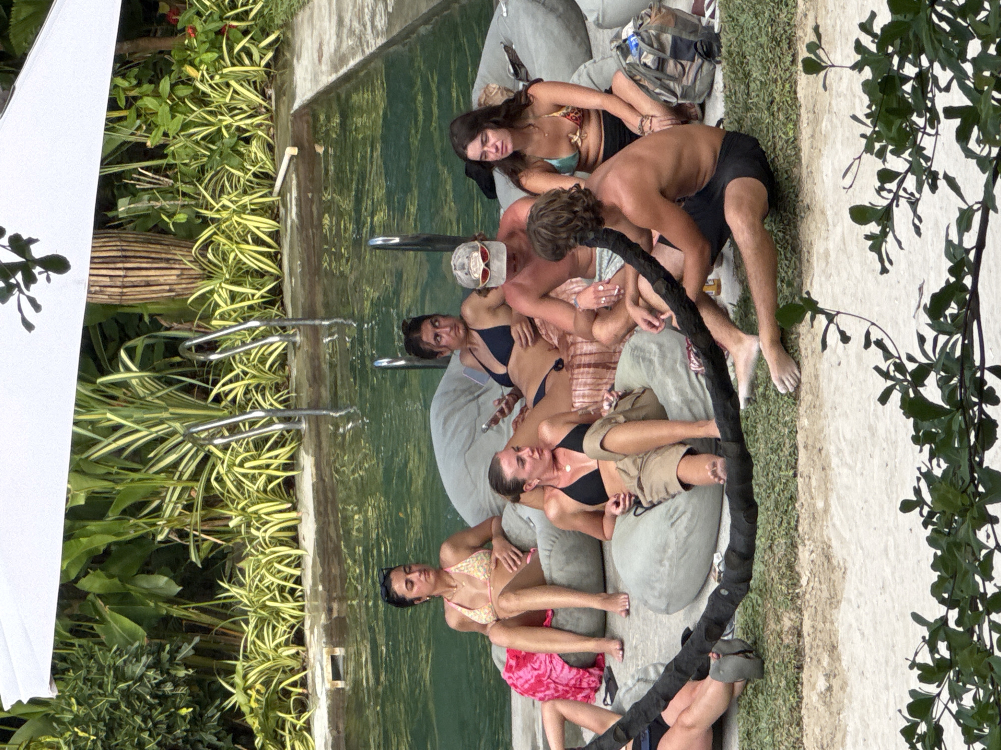 The image depicts a group of people lounging by a pool in General Luna, Philippines. There are approximately seven people visible, mostly women, dressed in swimwear or casual clothing. They are gathered on light-colored beanbag chairs or cushions placed adjacent to a pool of water. The pool is surrounded by greenery. A dark-colored cable or rope runs along the edge of the seating area. The background includes tropical vegetation.
