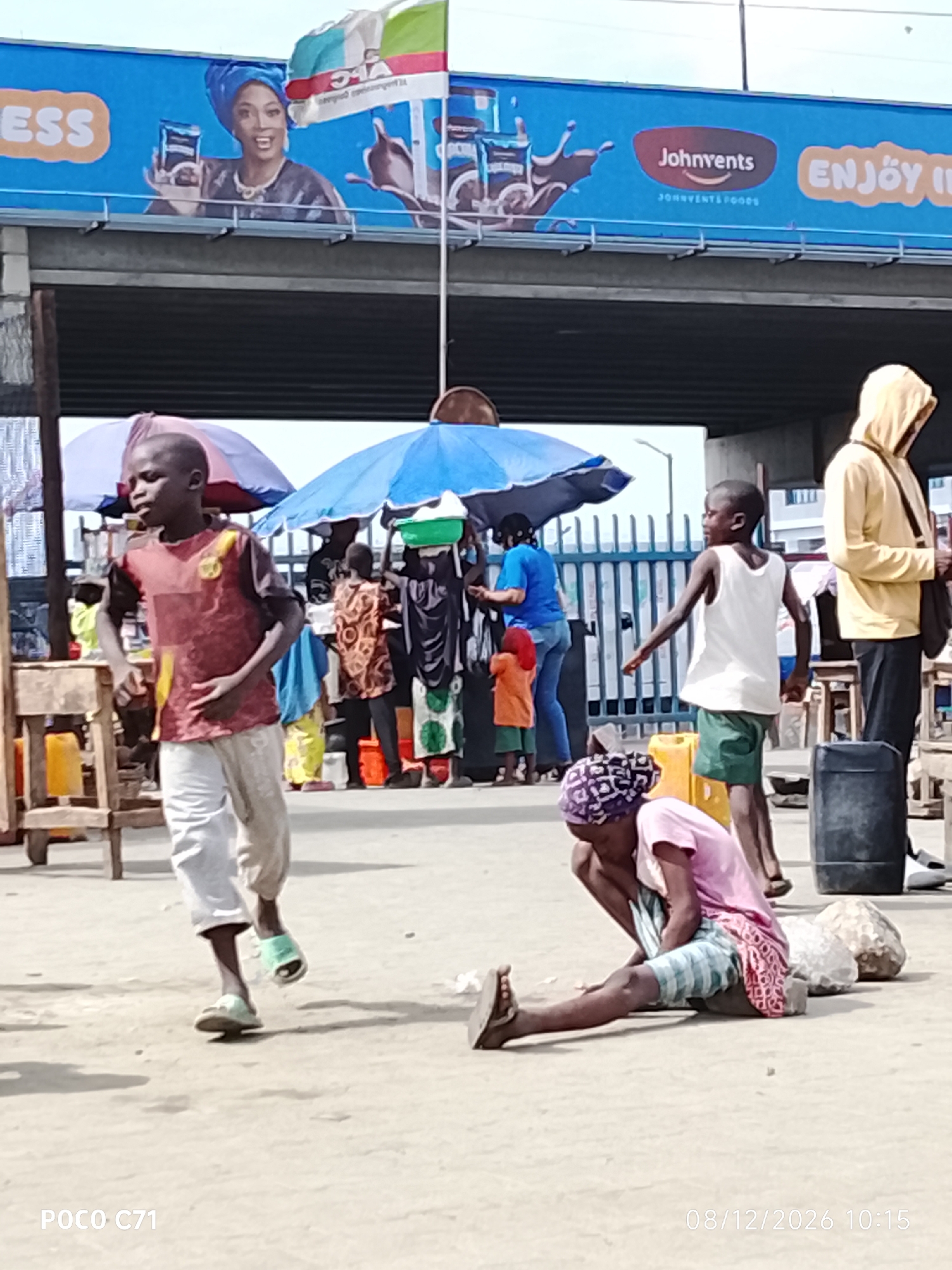 This image depicts a bustling outdoor scene, likely in Lekki, Nigeria, with a diverse group of people and various objects under a highway overpass. In the foreground, a young boy in a red and yellow patterned shirt and light-colored pants is walking, with green sandals on his feet. To his right, a young person in a pink shirt and checkered shorts is sitting on the ground, leaning on a stone, with their head bowed.
Behind them, several other individuals are visible. A person in a white tank top and green shorts is standing, looking to the left. Another person in a yellow hooded jacket is standing further back, facing away from the camera. Other figures, some obscured by umbrellas, are present in the middle ground, engaged in activities that suggest a marketplace or transit area.
Umbrellas, some blue and some patterned, are set up, indicating shelter from the sun. Yellow plastic containers and large black drums are scattered around the scene, suggesting commerce or storage. A wooden cart with yellow elements is to the left of the image.
Above the overpass, a large billboard displays advertisements. One section features a woman in a blue headwrap promoting a product. Another section shows the "Johnvents" logo and the word "ENJOY." A flag with green, white, and blue colors is also visible attached to a pole above the billboard. The ground surface appears to be a mix of sand and pavement. The overall impression is one of a vibrant, active urban environment.