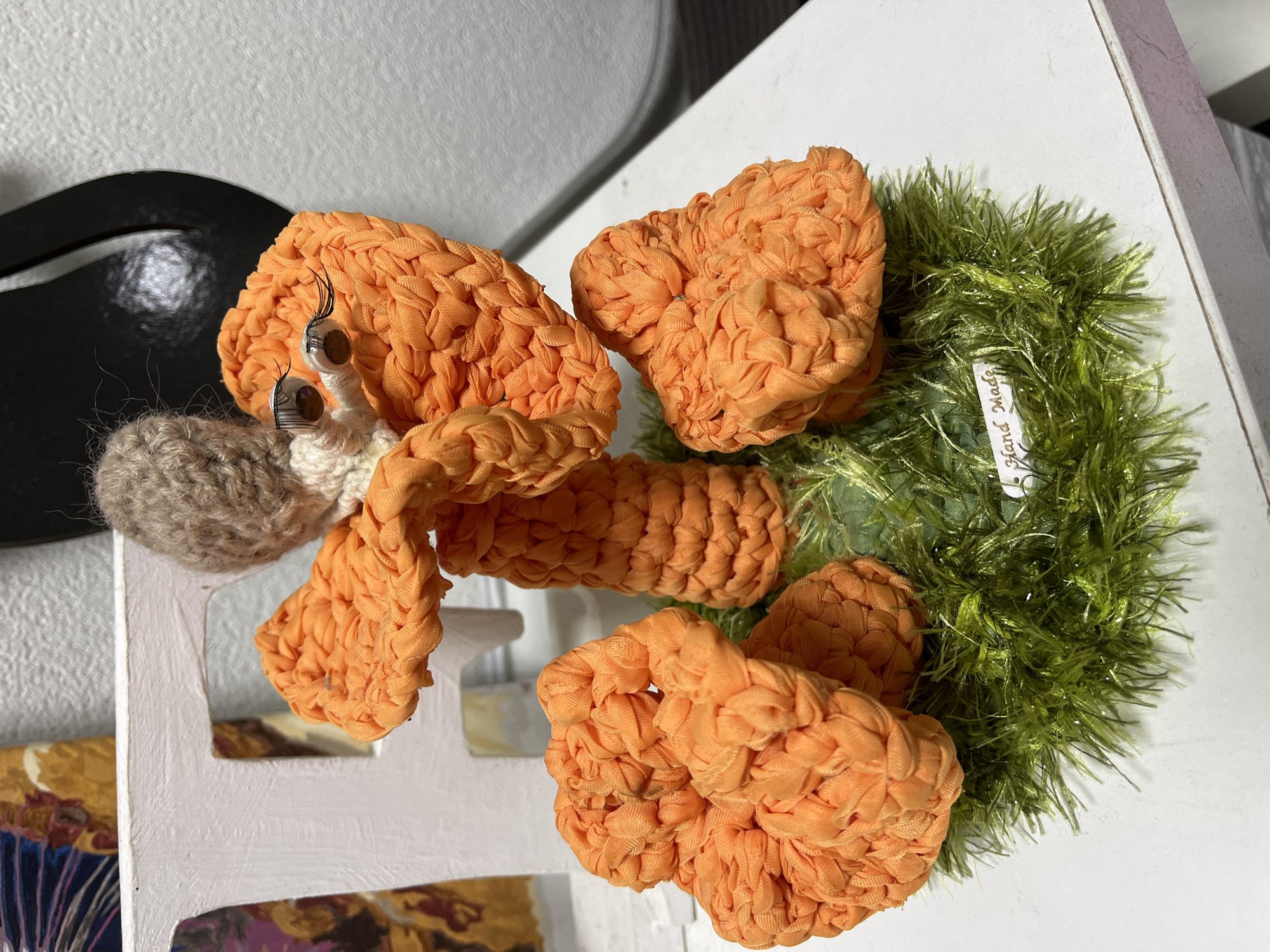 The image displays a handmade crochet craft, resembling a character with an orange body and a brown head, adorned with googly eyes and eyelashes. The craft is placed on a base of green, sparkly material, mimicking grass, with a small "Hand Made" tag visible. In the background, a white, distressed wooden structure, possibly a part of a chair or a shelf, is partially visible. To the right, a portion of a black, curved object is present. The scene is set indoors on a white surface, likely a table, with a textured white wall in the background. There is no indication of the location being in the city of Hrodna, Belarus.