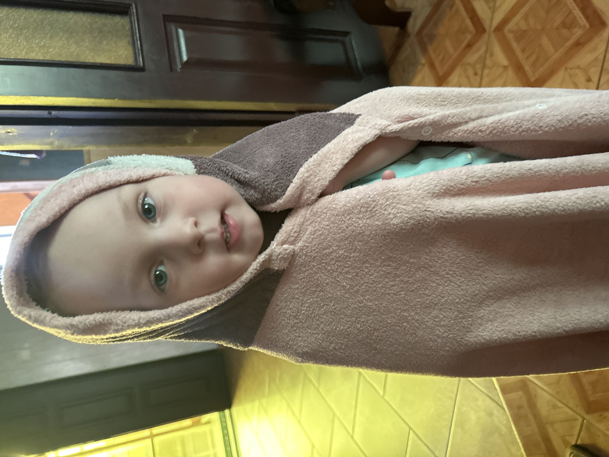 A young child is wrapped in a pink and brown hooded towel, standing in what appears to be a domestic setting. The child's face is visible, with wide green eyes and a slight smile. The background includes a doorway with a dark wooden door and a tiled floor. The lighting suggests an indoor environment, possibly a home. There is no information in the image to indicate the location is Gelendzhik, Russia.