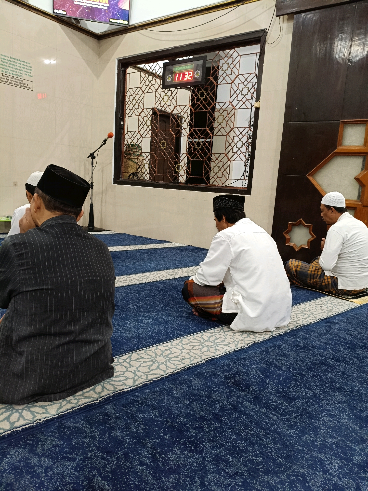 The image depicts an interior view of a mosque in Maguwoharjo, Indonesia. Three men are seen seated on a blue carpet with intricate white patterns. The man in the foreground, facing away from the camera, wears a black cap and a dark pinstriped shirt. To his right, another man, also facing away, is dressed in a white shirt and a patterned sarong. Further to the right, a third man, facing towards the left, is in a white shirt and sarong, with his back partially visible.
Behind the men, a large window with an ornate geometric screen is visible. Through the screen, a doorway can be seen, and to the left of the window, a digital clock displays "11:32". A microphone on a stand is positioned to the left of the window. The walls are a light color, and a portion of a television screen with colorful imagery is visible in the upper left corner. The overall environment suggests a prayer or congregational gathering in progress within the mosque.