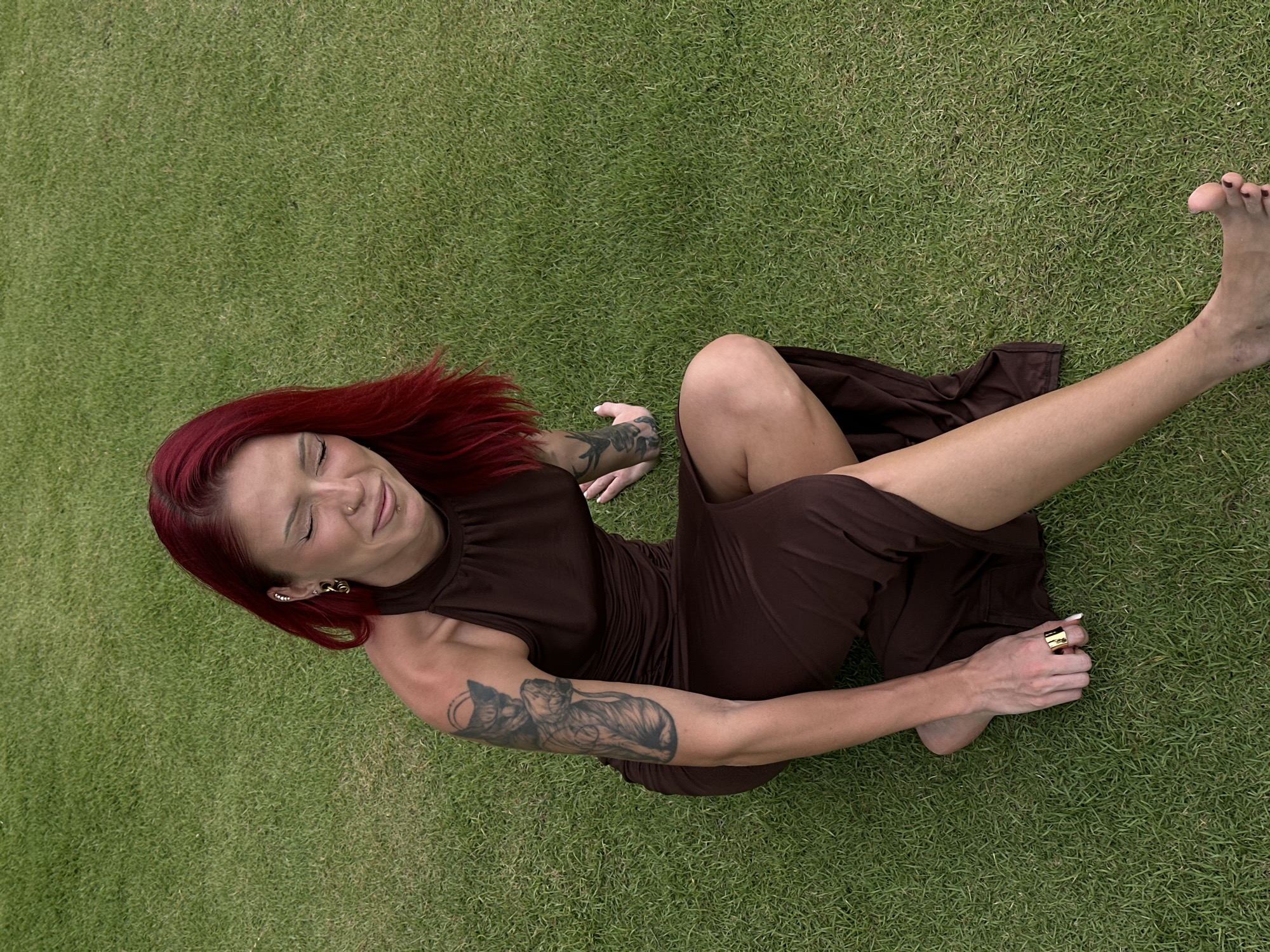 A woman with red hair, light skin, and tattoos on her arms is lying on green grass. She is wearing a brown dress and gold rings on her fingers and earrings. Her eyes are closed, and she appears to be smiling. The photograph appears to be taken from an aerial perspective. The location cannot be determined from the image alone, though the prompt indicates Nusa Dua, Indonesia.