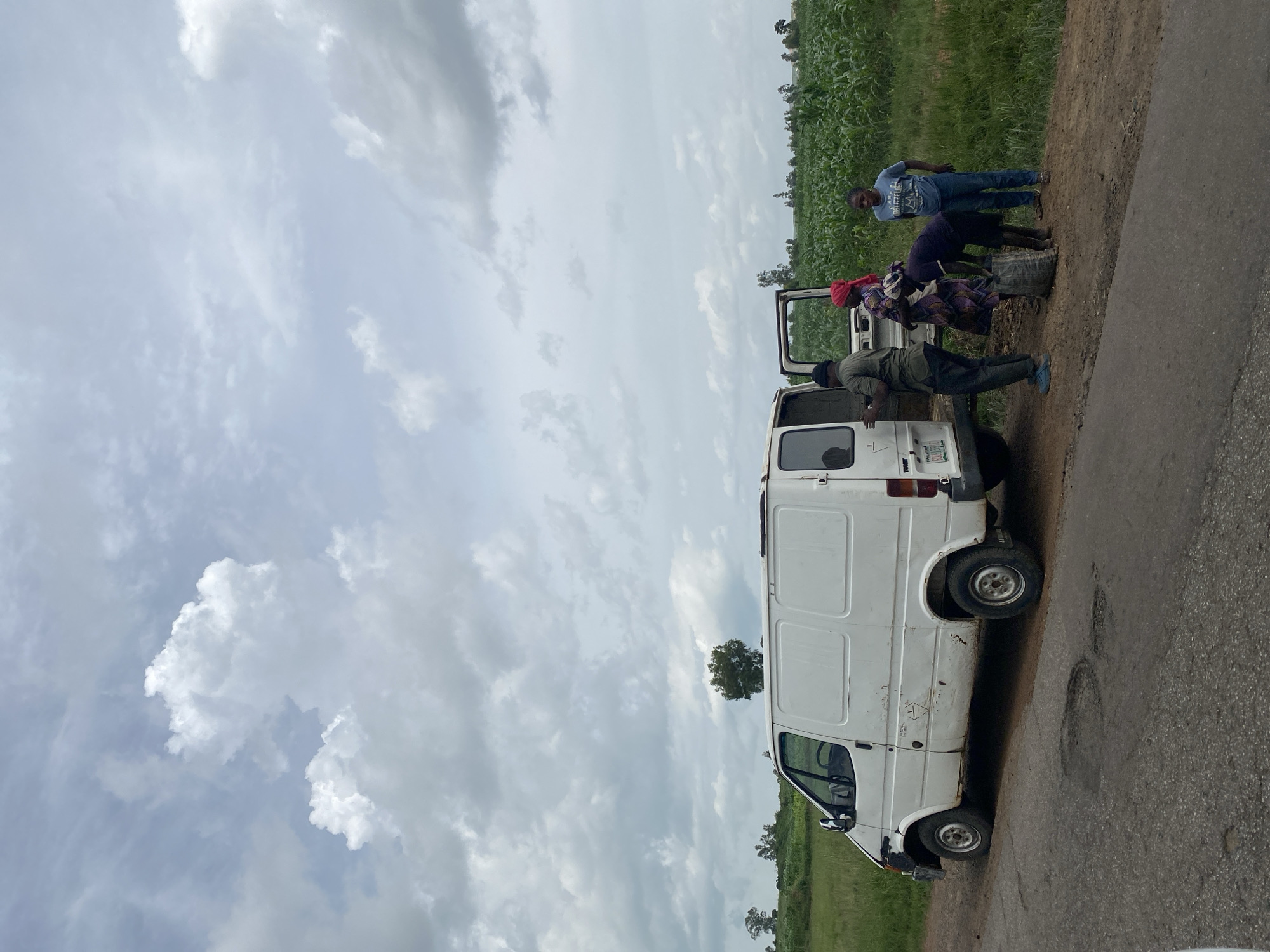 The image depicts a rural scene in Kuru, Nigeria, under a cloudy sky. A white van is parked on the side of a cracked asphalt road, which runs alongside a dirt path bordering a field of tall green crops, likely corn. Several people are gathered around the open rear doors of the van. One person, wearing a blue shirt and jeans, stands to the right, observing. Another individual, dressed in a purple and white patterned garment and a red headscarf, appears to be interacting with the van. A third person, wearing a dark shirt and shorts, is bending over, handling a large, dark-colored bag. A fourth person, in a green shirt and dark pants, is positioned near the van's interior. The van itself shows signs of wear and tear, with visible rust and damage to its exterior. In the background, a line of trees and shrubs stretches across the horizon, with more distant foliage suggesting a rural or agricultural setting.