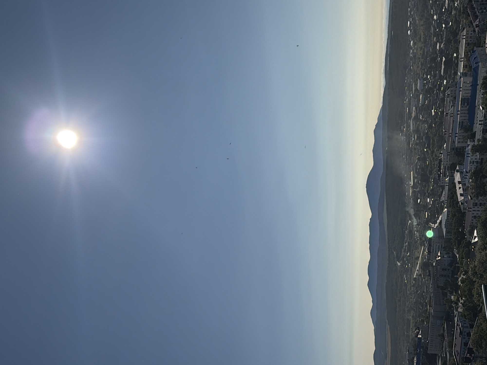 This is a photograph taken from a high vantage point, looking out over a city and a range of distant mountains. The sky is a clear, bright blue, with the sun prominently visible in the upper portion of the frame, casting a strong glare and lens flare. The sun is a bright white circle with a halo of light around it, suggesting it is high in the sky, likely mid-morning or afternoon.
Below the sky is a hazy horizon where the sky transitions into a lighter blue and then a pale white, possibly indicating a layer of distant atmospheric haze or fog. Beneath this haze lies a dark, undulating silhouette of mountains. These mountains appear to be covered in vegetation and are largely devoid of any visible human structures, adding a natural backdrop to the urban landscape.
The foreground of the image is dominated by a city. The buildings are primarily low to mid-rise, with a mix of apartment blocks and smaller structures. The architecture suggests a post-Soviet era, with many buildings featuring uniformly colored facades and balconies. Greenery, in the form of trees and shrubs, is interspersed throughout the city, particularly in the lower portions of the image. Roads and pathways wind through the urban area. In the middle ground, some wisps of smoke are visible, possibly from chimneys or industrial activity.
There are several small, dark specks scattered across the sky, which appear to be birds in flight. One particularly bright green circular anomaly is visible on one of the buildings in the mid-ground, likely a lens flare or artifact from the camera. The overall lighting suggests it is a sunny day. The scene is a panoramic view of a city nestled between natural landscapes, captured under a bright, clear sky. The location is the city of Korsakov, Russia.