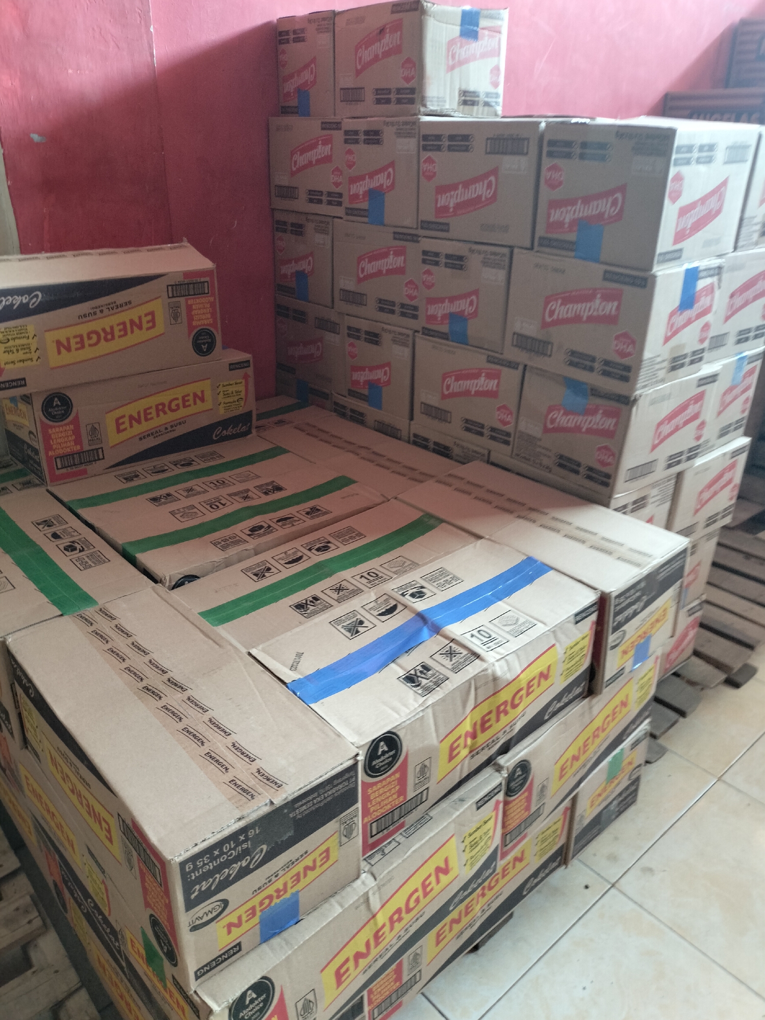 The image shows stacks of cardboard boxes of Energen cereal and milk and Champion brand boxes in what appears to be a warehouse or storage area. The boxes are predominantly cardboard-colored with red and yellow branding. Some boxes have blue tape sealing them. The environment suggests a commercial setting, likely a distribution point or retail storage. The floor is tiled, and a pink wall is visible in the background. No people are present in the image. The location of Langsa, Indonesia, is not explicitly depicted or implied by visual cues within the image itself, other than the presence of Indonesian language text on the boxes.