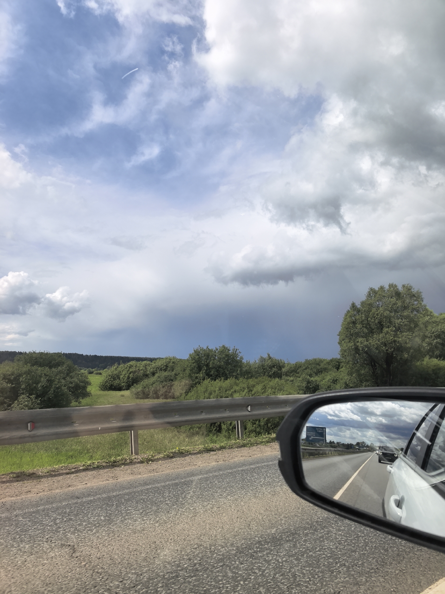 The image shows a view from inside a vehicle traveling on a road, possibly near Chernovskoe, Russia. The sky is overcast with a mix of blue and gray clouds; contrails are visible. A dark patch of precipitation is visible in the distance. A guardrail runs along the side of the road. Beyond the guardrail, a field of low-lying green vegetation is visible. The car's side mirror shows a reflection of the road behind, including other vehicles and a road sign. The reflection shows the road continuing straight. The road surface is asphalt.