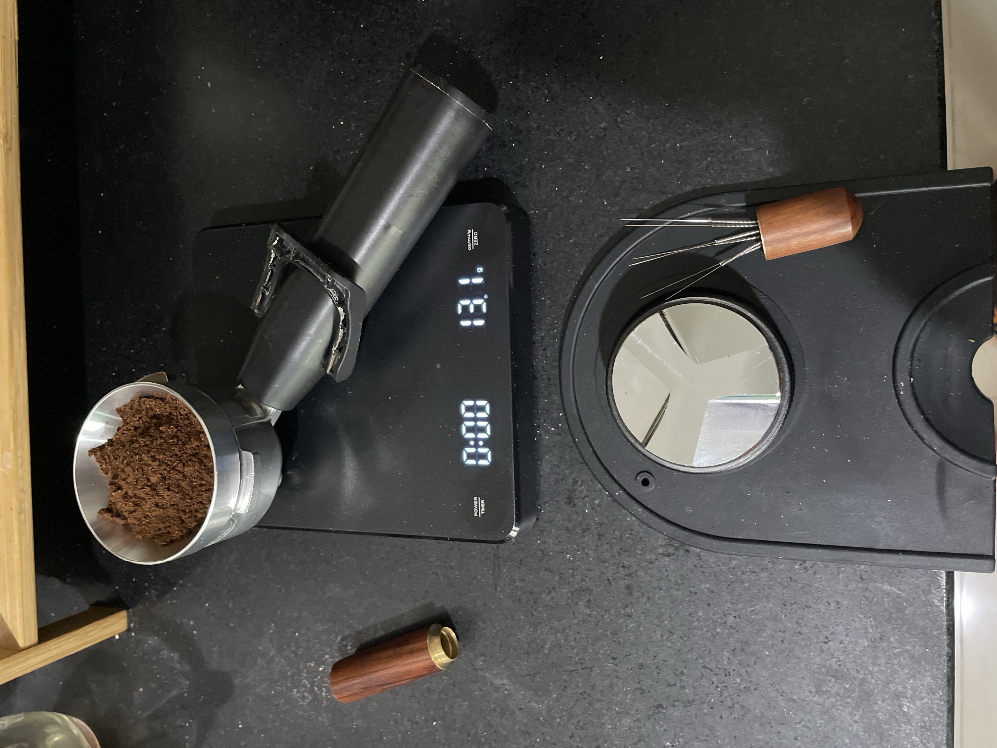 The scene appears to be a countertop setup for preparing espresso.
Objects present include:
* A portafilter filled with ground coffee, resting on a digital scale. The scale reads "13.1g" and "0:00"
* A coffee distribution tool with fine needles and a wooden handle, and its protective wooden case.
* A black espresso machine with two circular platforms.
The countertop is black with a fine-grained texture. A blurred wooden structure is present in the lower left corner.
There are no people visible in the image. The scene depicts an espresso-making preparation process. There are no elements that suggest Kuta Selatan, Indonesia.