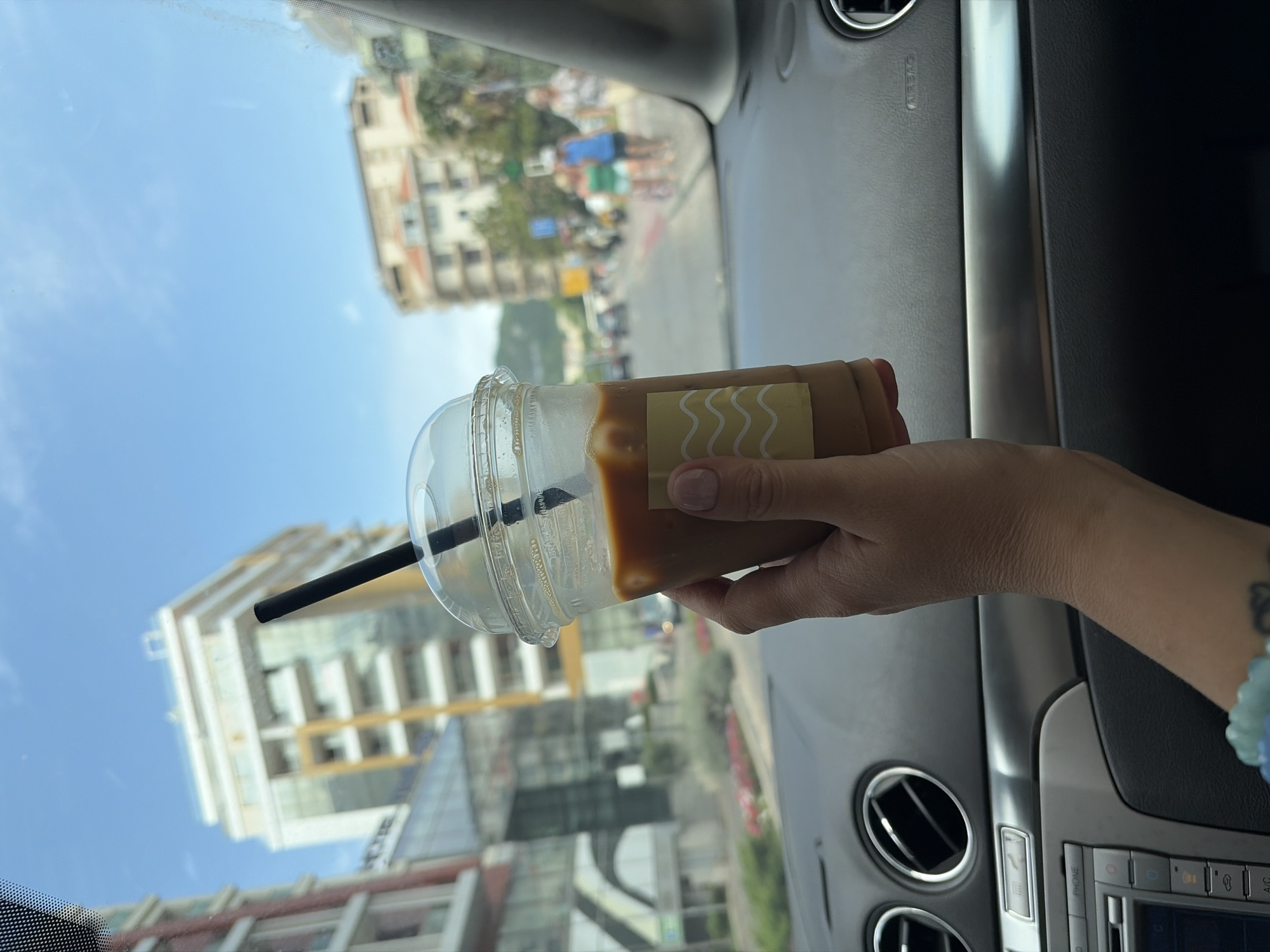 The image presents a first-person view from inside a car, looking out the passenger window on a bright, sunny day in Budva, Montenegro. In the foreground, a hand holds a clear plastic cup of iced coffee. The beverage is a light brown color, and a black straw extends from the domed lid. A light yellow label with white wavy lines is affixed to the cup. A light blue and white beaded bracelet is visible on the wrist of the hand holding the drink.
The car's interior is partially visible on the right and lower parts of the frame, showing a grey dashboard with circular air vents and a silver trim piece. The word "AIRBAG" is faintly legible on the dashboard above one of the vents.
Outside the car, the background reveals a vibrant blue sky with minimal cloud cover. Several multi-story buildings, featuring both modern and more traditional architecture, line a street. Trees and other vehicles are present, along with blurred figures of people suggesting typical urban activity. The strong daylight indicates it is daytime. The scene captures a casual moment, possibly during travel or a stop, with a refreshing drink and a view of the city.
