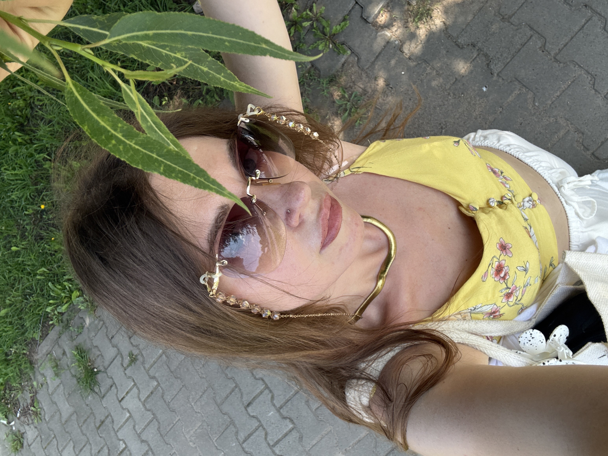 A young woman is taking a selfie outdoors. She is wearing sunglasses, a yellow floral top, a gold necklace, and white shorts. She is holding a leafy branch over her head with her right arm. The background shows a paved walkway and grass. The scene appears to be in a park or a residential area.