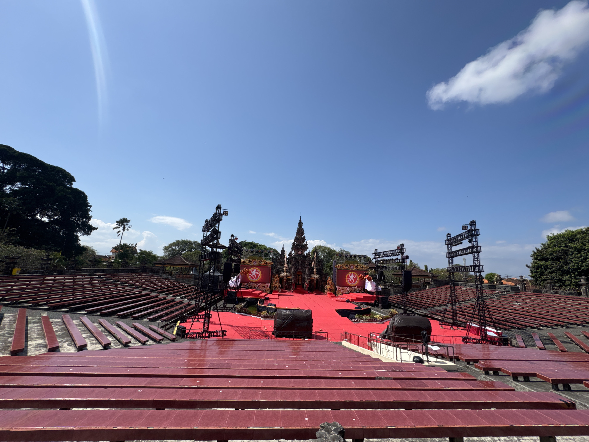 An outdoor amphitheater is set up for a performance. The seating is arranged in multiple tiers of dark red benches. In the center of the amphitheater, a vibrant red carpet covers the performance area. Several elaborate structures, resembling traditional Balinese architecture, are positioned on the stage, adorned with gold accents and large circular banners featuring a lion-like creature. Black tarps cover large objects in front of the stage. Tall black metal structures, likely for lighting and sound equipment, are erected on either side of the performance area. The scene is bathed in bright sunlight under a clear blue sky with a few scattered clouds. Lush green trees and vegetation surround the amphitheater. The location is the Ardha Candra open-air amphitheater in Denpasar, Bali, Indonesia. There are no people visible in the image.